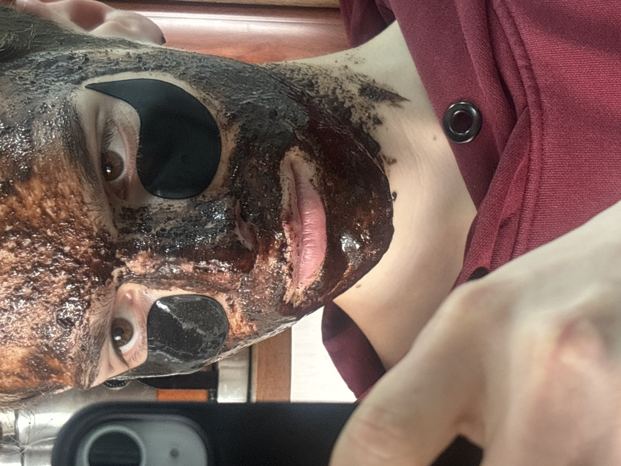 A close-up shot focuses on a person's face covered in a dark, textured substance. Black eye patches are placed under each eye. The person has fair skin, brown eyes, and pink lips. A portion of their neck is visible, along with a dark red garment with a button or eyelet. A hand holding a smartphone is visible at the bottom of the frame. The background consists of wooden surfaces. There is no information to support a location in Gyumri, Armenia.