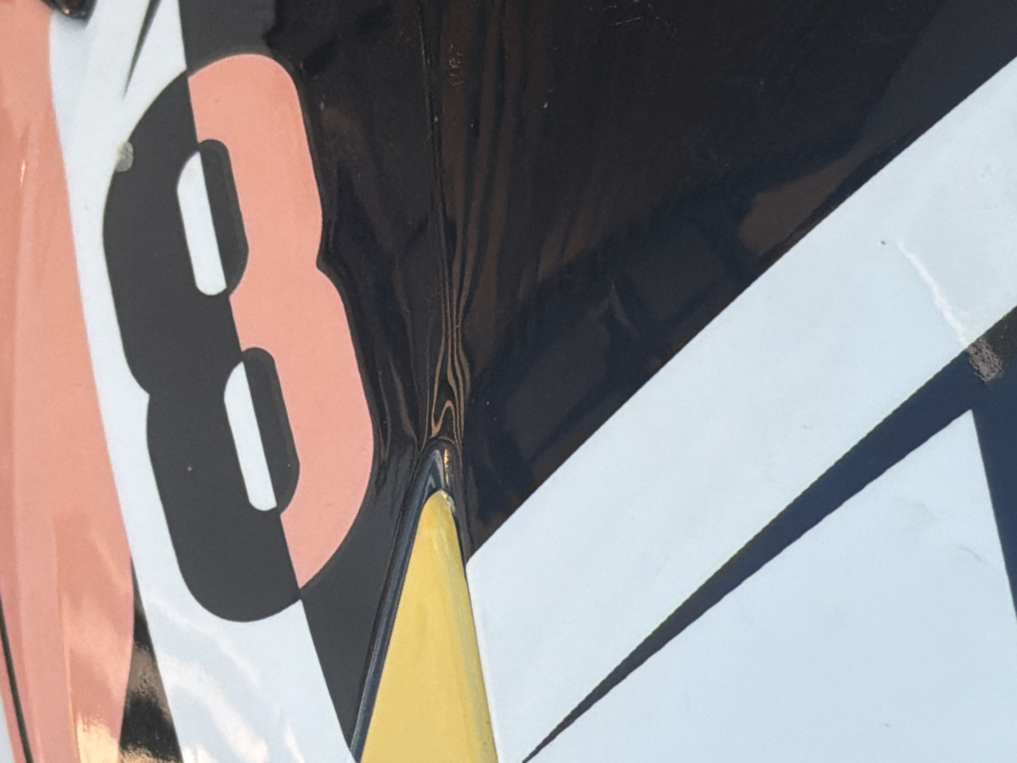 This is a close-up, high-angle shot of a section of a vehicle's exterior, possibly a car or boat, with a brightly colored graphic design. The design features a large, stylized black number "8" outlined in a light pink, set against a white background. The "8" has two oval-shaped cutouts. A section of yellow is visible to the left of the number, transitioning into a black area that appears to be the body of the vehicle. The surface is glossy and reflective, showing some minor scuffs and imperfections. The background consists of a peachy-pink color on the upper part of the image. The scene is devoid of any people or activity, and the specific location in Baan Chaweng, Thailand, cannot be determined from the visual information provided.