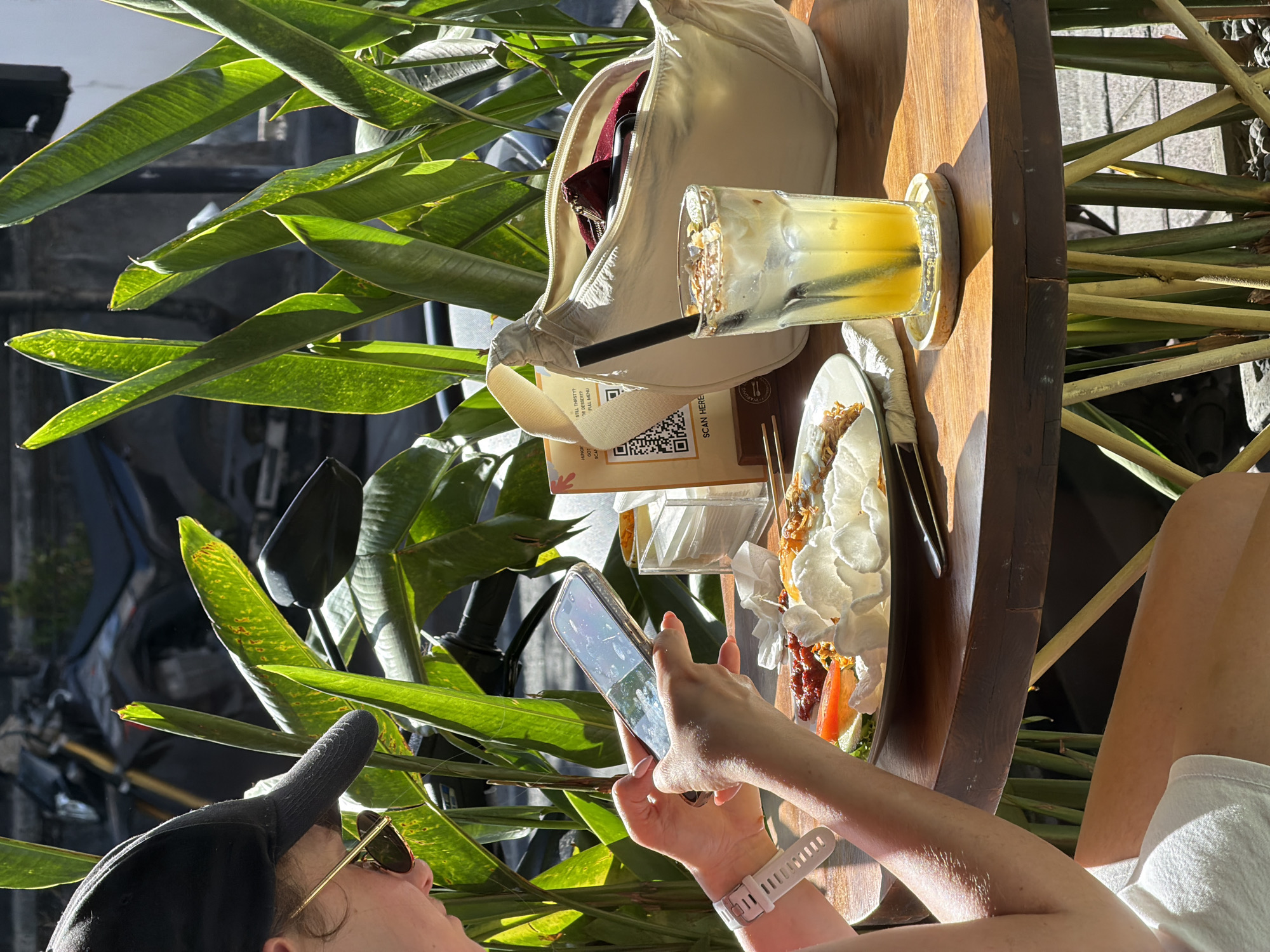 A person is using a smartphone to take a photo of a meal and drink on a wooden table in North Kuta, Indonesia. The table is surrounded by lush green plants. A white bag sits next to the drink. The person is wearing a white fitness tracker, a black baseball cap, and sunglasses. A car is visible in the background.