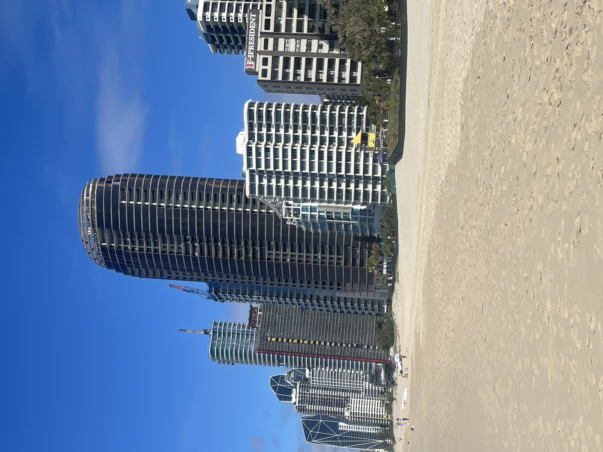 A wide shot captures a sunny day at a beach in the Gold Coast, Australia, with a clear blue sky and a few clouds. The foreground is dominated by a sandy beach. In the midground, a line of modern high-rise buildings forms the city skyline. Among them, a tall, dark, curved building stands out, possibly under construction with exposed floors and a skeleton-like top. Adjacent to it are several other apartment buildings with white facades and balconies. A construction crane is visible near one of the buildings. A sign reading "PRESIDENT" is mounted on one of the buildings. In the distance, palm trees and other greenery are visible between the buildings. A few people are scattered on the beach, some walking and others near a parked white vehicle and a surfboard. The ocean is not visible in this frame.