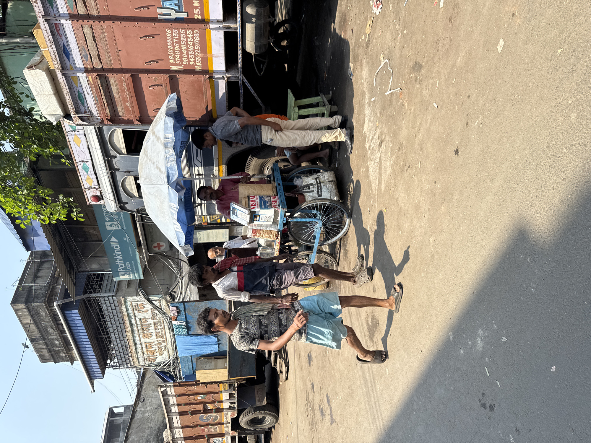 The image shows a street scene in Kolkata, India. A man is standing in the foreground wearing a patterned shirt and blue shorts, walking to the right of the frame. Behind him, two men stand near a mobile food cart with a blue umbrella. One of the men is wearing a red shirt and attending to the cart. Another man in a gray shirt is leaning against a large truck with a red and brown cargo area. The truck bears a sign with phone numbers in Bengali. Buildings are visible in the background, including a building with the sign "Pathkind Labs."