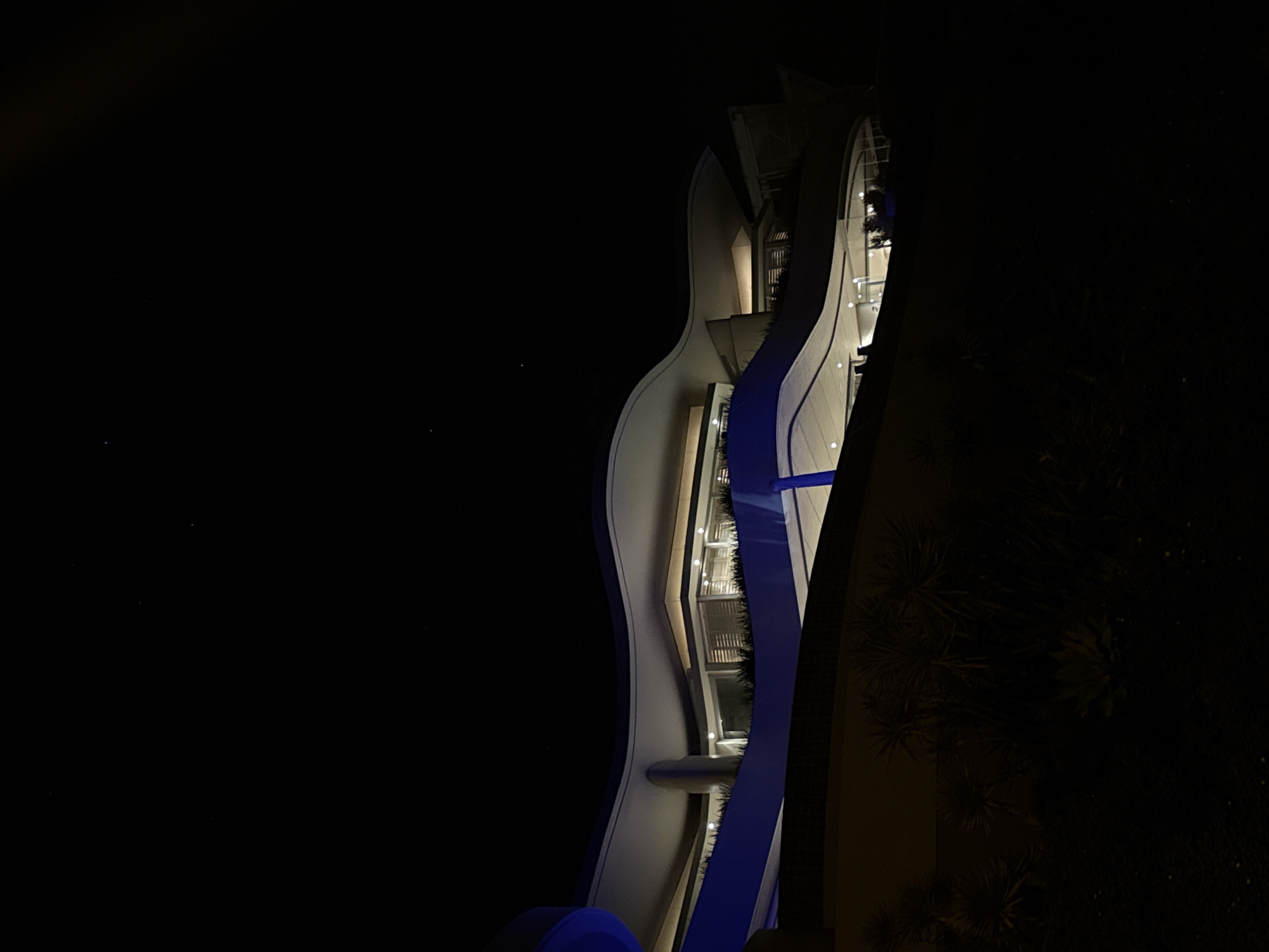 The image depicts a modern building at night. The building has a unique, curved design and is illuminated with white and blue lights. It appears to have balconies or terraces with greenery visible behind the glass windows. The surrounding environment is dark, with some vegetation visible at the base of the building. The location is Cape Town, South Africa, according to the metadata. There are no people visible in the image.
