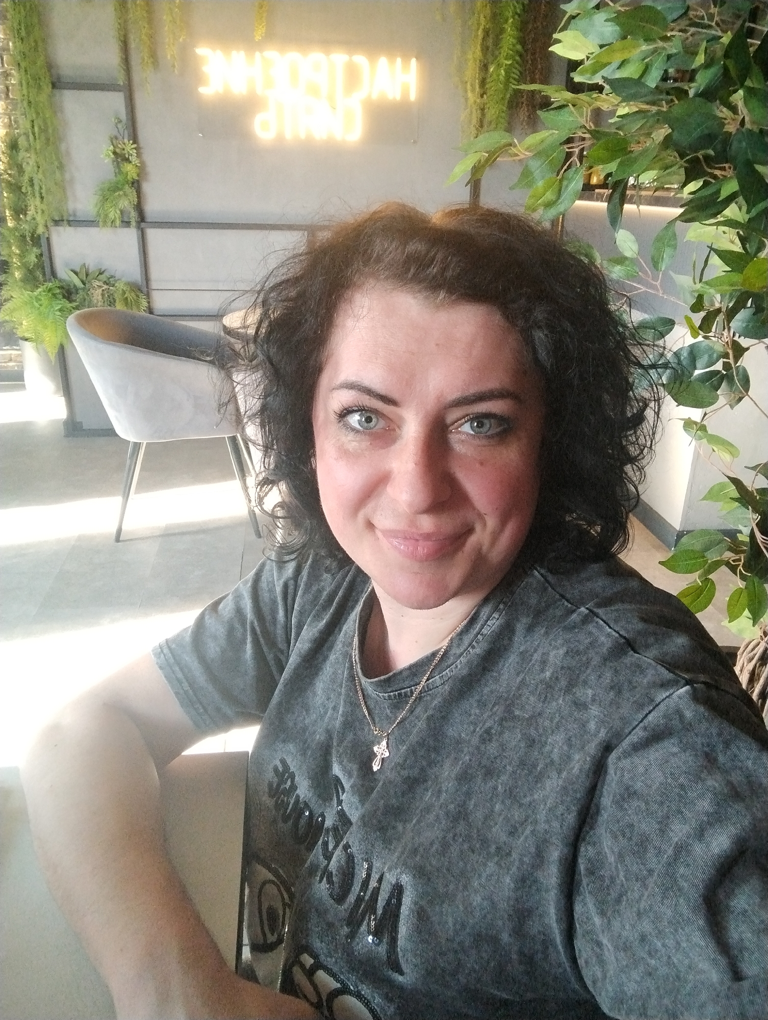 A medium shot captures a woman indoors. She has fair skin, blue eyes, and dark curly hair. She is wearing a grey t-shirt and a silver necklace with a cross pendant. She is seated, with her left arm resting on the arm of the chair.
In the background, there is a grey chair, greenery, and a neon sign. The location is identifiable as being within the city of Votkinsk, Russia.