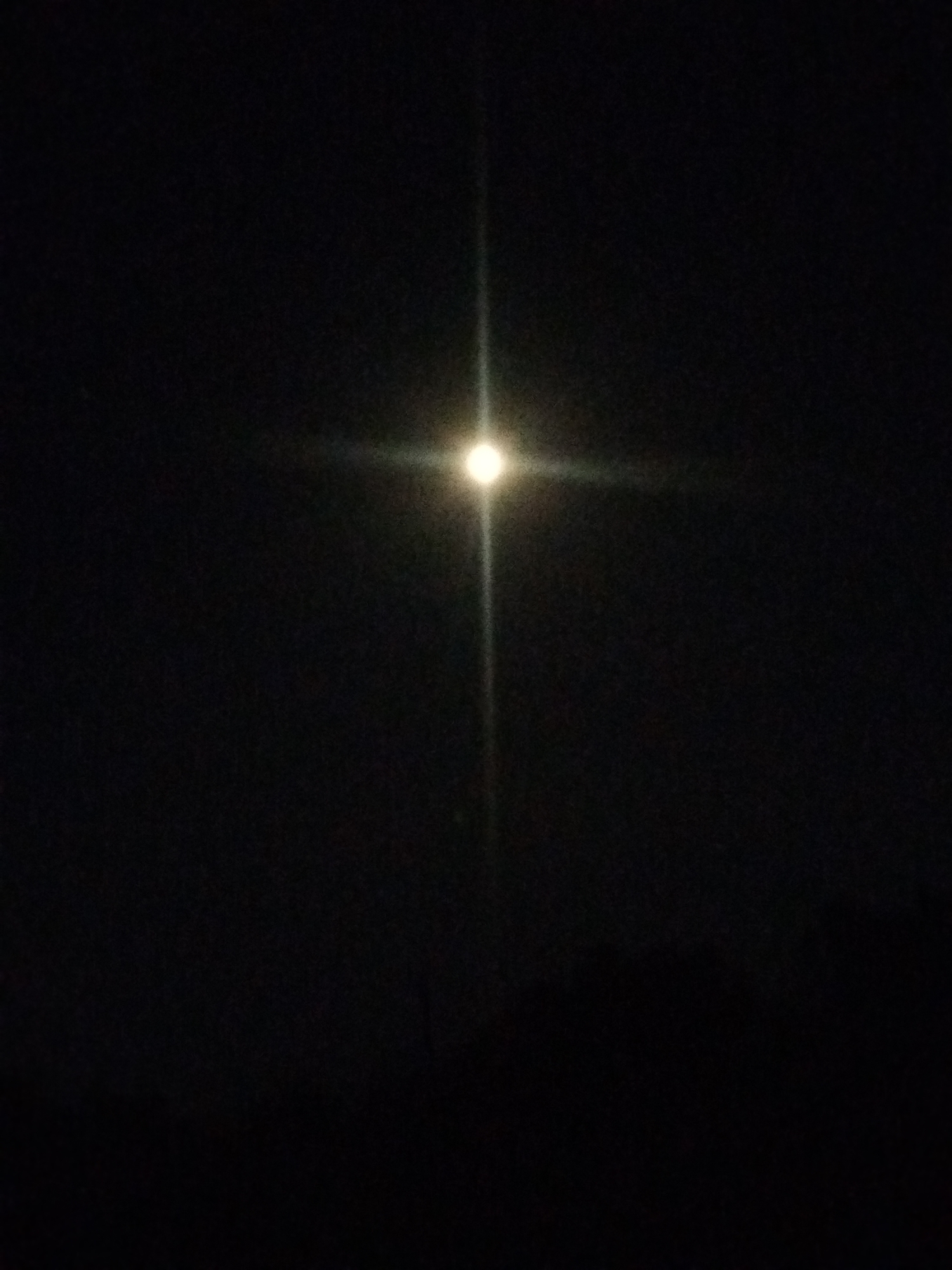 The image displays a dark, night sky with a bright, glowing object in the center, emitting a starburst effect. The object is likely the moon, heavily overexposed by the camera. Faint horizontal and vertical lines of light extend from the central glow, creating a cross-like appearance. The lower portion of the image shows a dark, indistinct horizon line, suggesting the presence of trees or buildings, but these are obscured by the darkness. The overall scene is characteristic of a nighttime photograph taken with a light source that is too bright for the camera's exposure settings. There are no people or discernible objects other than the celestial body and the faint silhouette of the landscape. The location is Kostiantynivka, Ukraine.