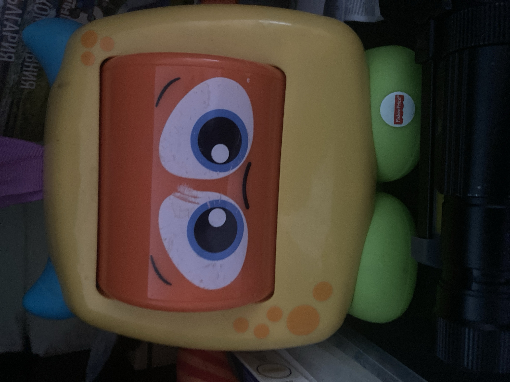 The image shows a close-up, eye-level, frontal view of a yellow toy with a cartoonish face, featuring large blue and white eyes with black pupils and a downturned mouth, giving it a sad expression. The toy has two blue horns on top and orange polka dots on its sides. Its lower half consists of two green, bulbous feet. A small circular white sticker with the "Fisher-Price" logo is visible on the right green foot. The toy is positioned in what appears to be an indoor setting, possibly a storage area, with dark backgrounds and some out-of-focus objects visible. There is no indication of the city of Burgas, Bulgaria, in the image.