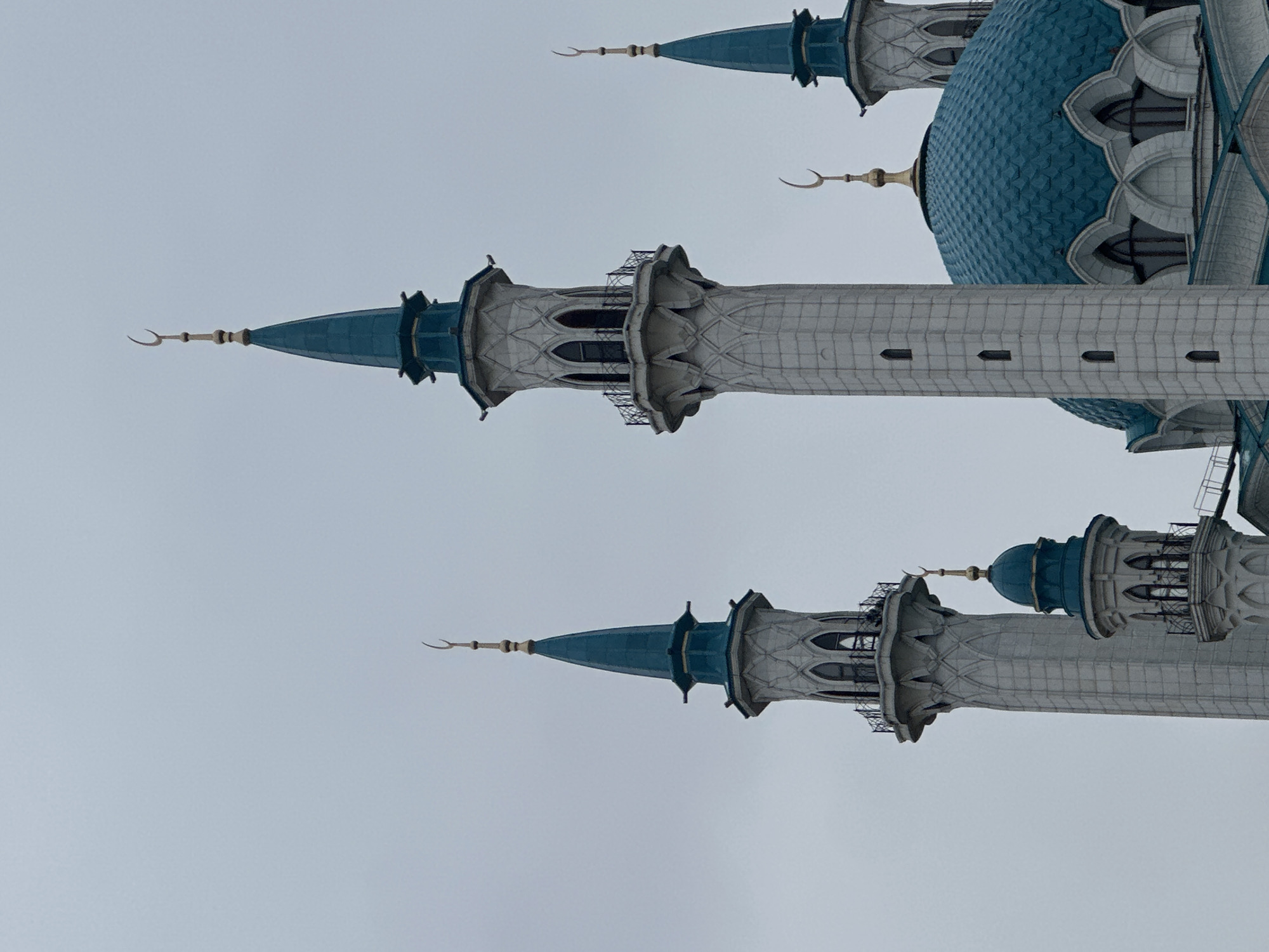 The image shows a view of the Qolsharif Mosque in Kazan, Russia. Visible are multiple minarets, with blue conical tops and crescent moon finials, as well as part of the larger central blue dome. The minarets are pale gray, with ornate details. The sky is overcast. No people are visible in the frame. The mosque is situated against a backdrop of a uniform gray sky.