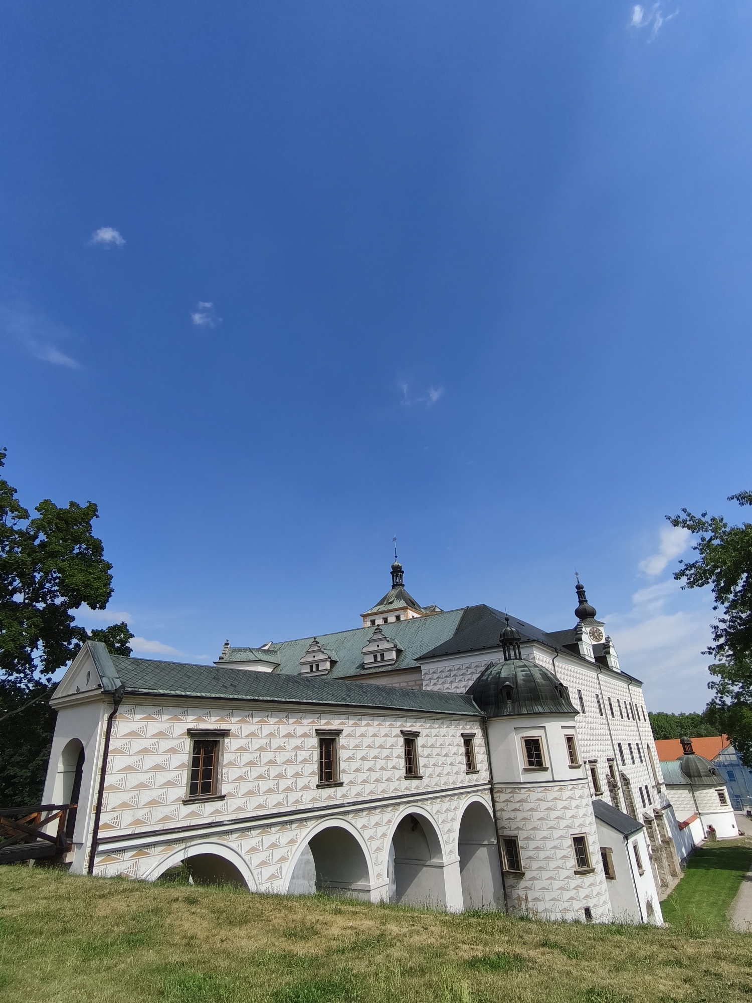 The image shows the exterior of Pardubice Castle, located in Pardubice, Czechia. The castle features white walls adorned with a repeating geometric pattern of triangles and lines in shades of beige and gray. Several sections of the castle are visible, including a building with arched openings on the ground level and windows on the upper floor. This section is connected to a larger structure with a dark-tiled roof and multiple towers, one of which has a clock face. The architecture suggests a historical building with Renaissance influences. The castle is situated on a grassy slope, with trees visible on the left and right sides of the frame. The sky above is a clear, vibrant blue with a few scattered white clouds. The lighting indicates it is daytime, likely a sunny afternoon. No people are visible in the image.