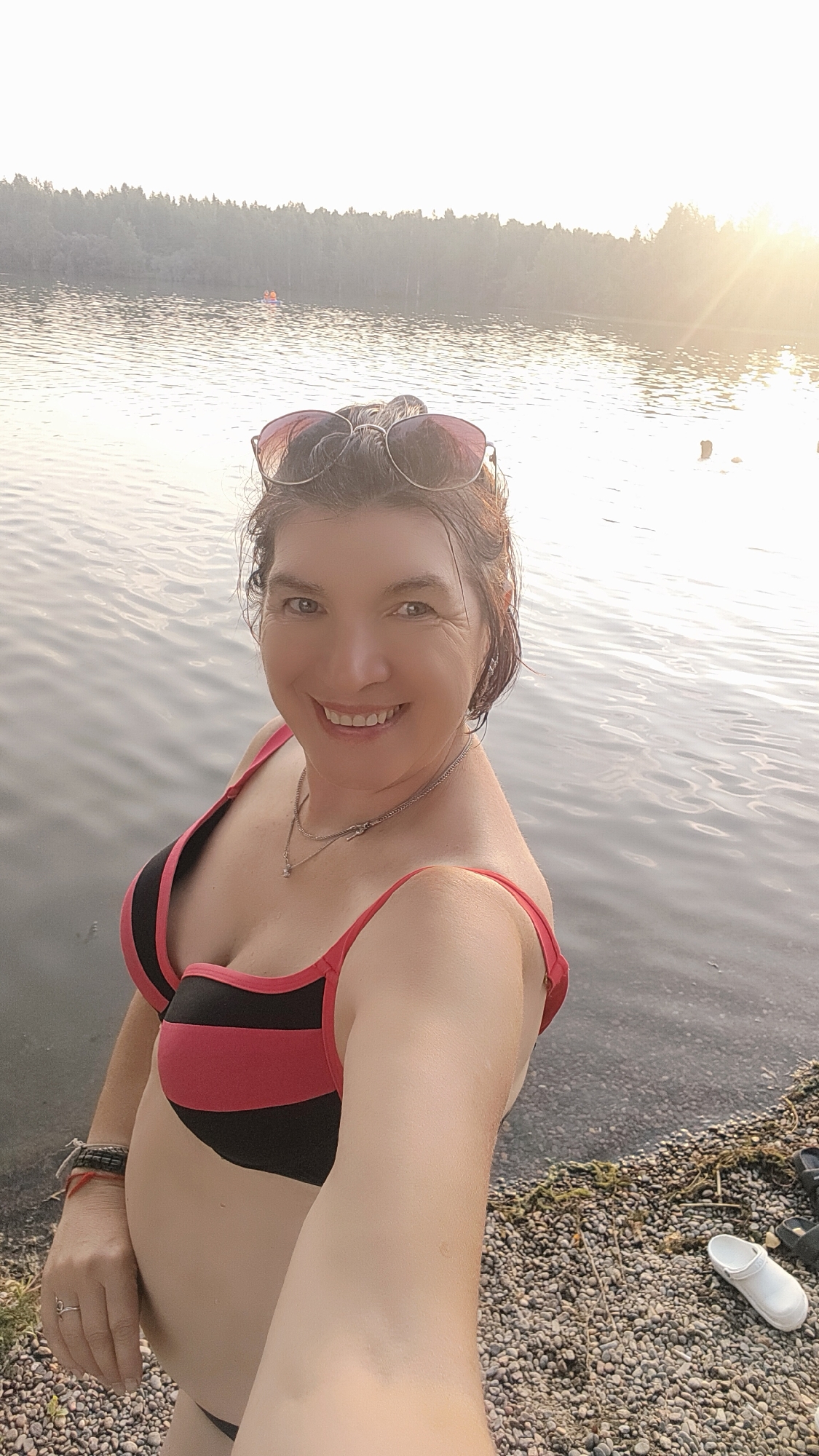 The image shows a woman in a bikini taking a selfie by a lake. She has short, dark hair, is wearing sunglasses on her head, and a necklace. The bikini is black and red striped. In the background, there is a forested shoreline and a hazy, sunlit sky, suggesting it is late afternoon or early evening. The water appears calm. The ground beside her is covered in small stones and some scattered vegetation. There are also a pair of white clogs visible on the shore. The woman is smiling broadly, conveying a happy and relaxed emotion. The scene appears to be set in Terent'yevo, Russia, likely during warmer weather given the attire. There is no visible text in the image.