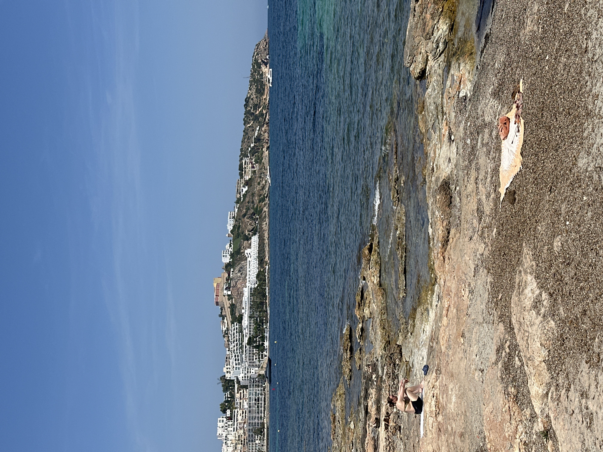 The photograph depicts a coastal scene. On the right, rocky terrain extends to the ocean's edge. A woman is seen sunbathing on a white towel. A beach bag is nearby. In the distance, the ocean stretches to the horizon, where the skyline is marked by buildings and hills. The sky is clear and blue. The scene is likely located in Ibiza, Spain, given the architectural style of the buildings and the coastal environment.
