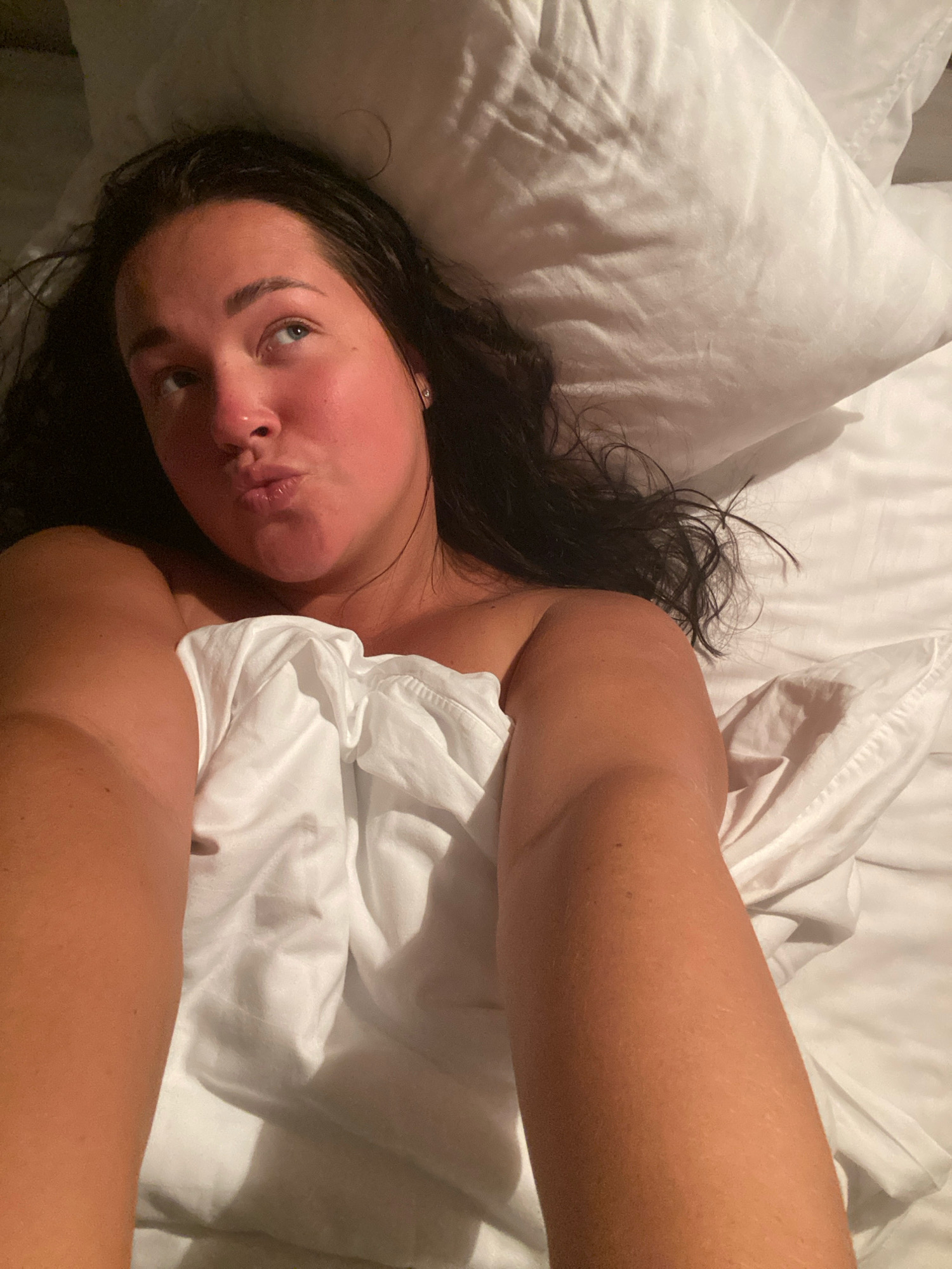 A woman is lying in bed, covered by a white duvet, with her head resting on a white pillow. She has long, dark hair and is looking upwards and to the left, with her lips pursed. Her skin appears flushed, particularly on her cheeks. The environment is consistent with a bedroom. No specific details about the location in Nebug, Russia are discernible from the image itself.