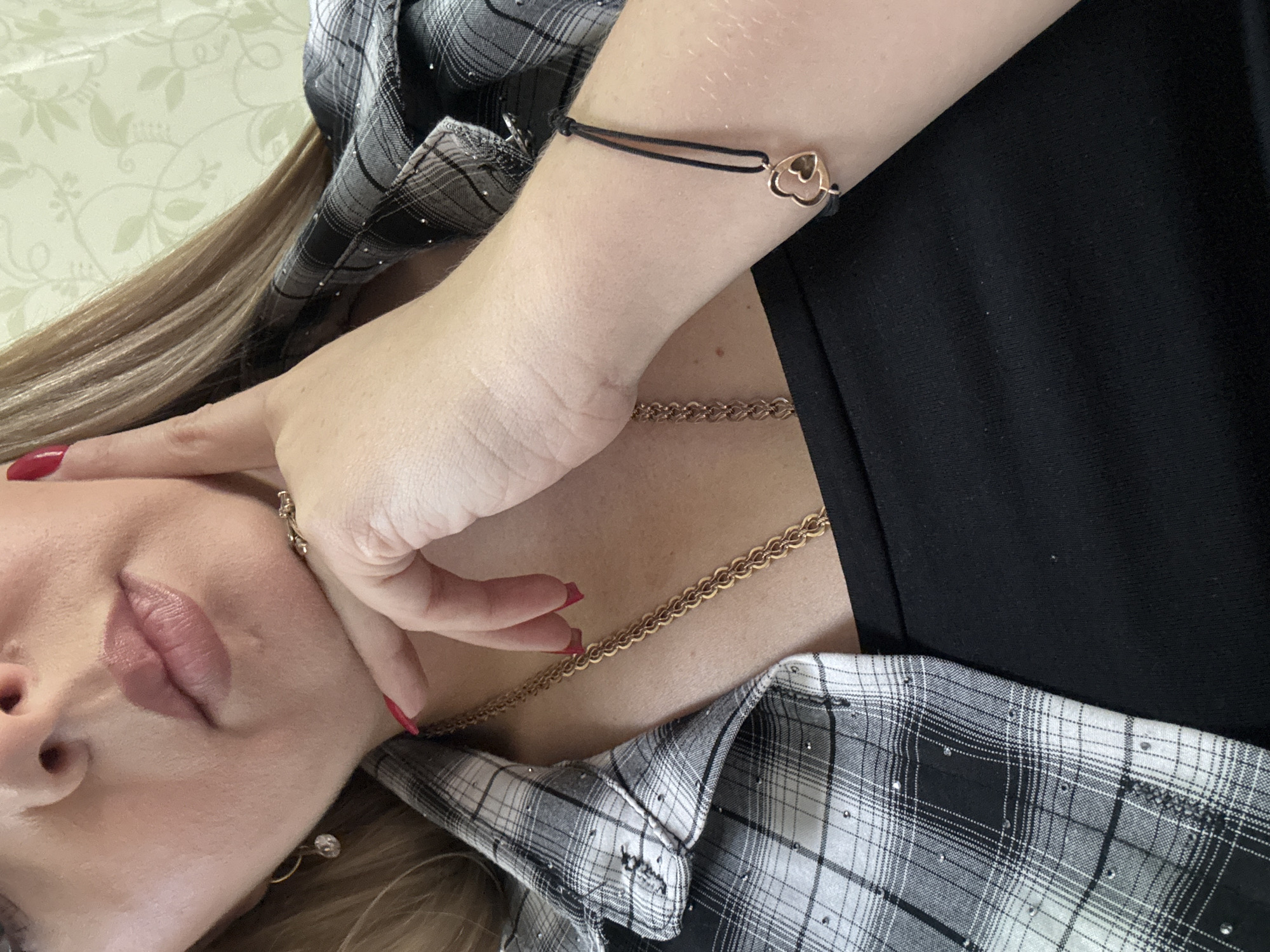 A medium shot captures a woman from the chest up, positioned in front of a subtly patterned wallpaper. She is wearing a black, sleeveless top and an open, grey and black plaid shirt adorned with small, reflective embellishments. Her right hand is raised, with index and middle fingers resting on her chin, her thumb supporting her jaw. Her nails are painted a vibrant red. A gold chain necklace is visible around her neck. On her wrist, she wears a thin black cord bracelet with a rose gold charm featuring two intertwined hearts. A single diamond earring dangles from her left ear. The lighting is even, and the background appears to be indoors.