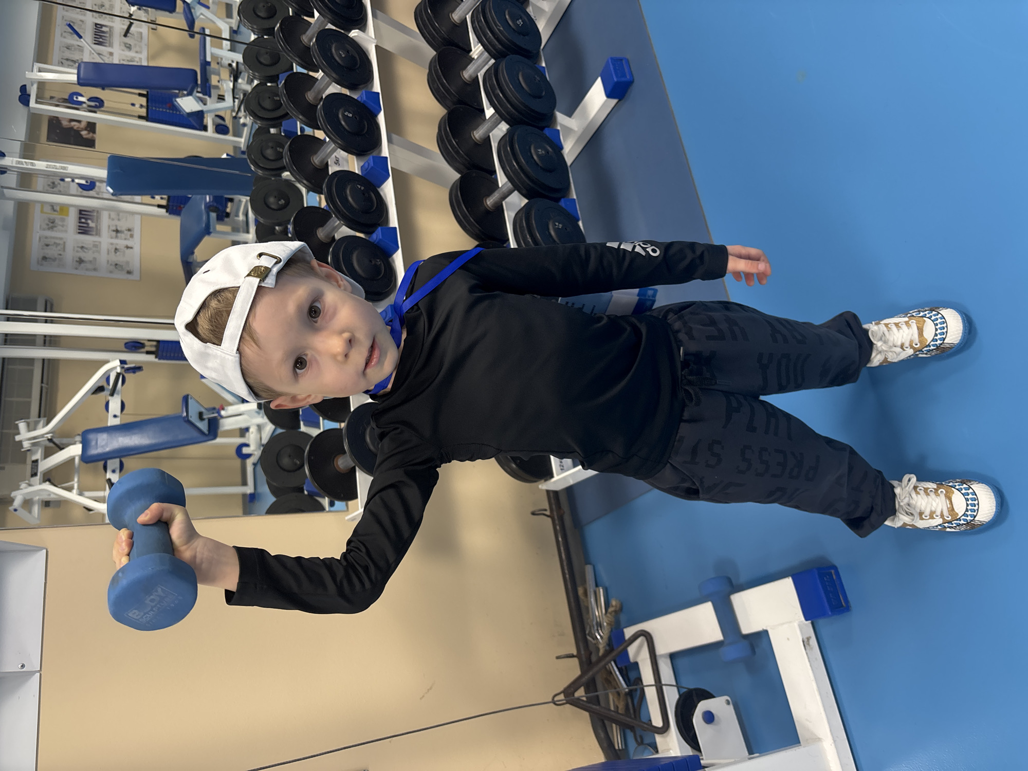The image shows a young boy in a gym in the city of Chaikovsky, Russia. He is wearing a black long-sleeved shirt, dark patterned sweatpants, and white sneakers. He also has a white baseball cap on his head. The boy is holding a blue dumbbell in his right hand. The gym has weight racks filled with black weights in the background. The floor is blue, and the walls are a light beige color. There are also various gym machines visible in the background.