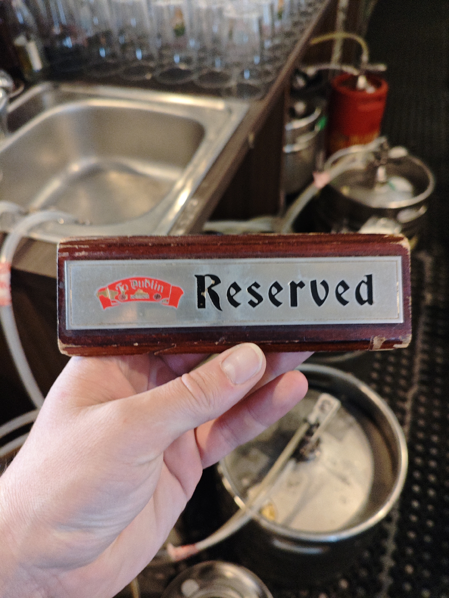 A person's hand is holding a wooden sign with a silver plaque that reads "To Dublin" on the left side and "Reserved" on the right side. The background shows a stainless steel sink, a red keg, and other bar equipment, suggesting a pub or restaurant setting. The location is not visible in the image.