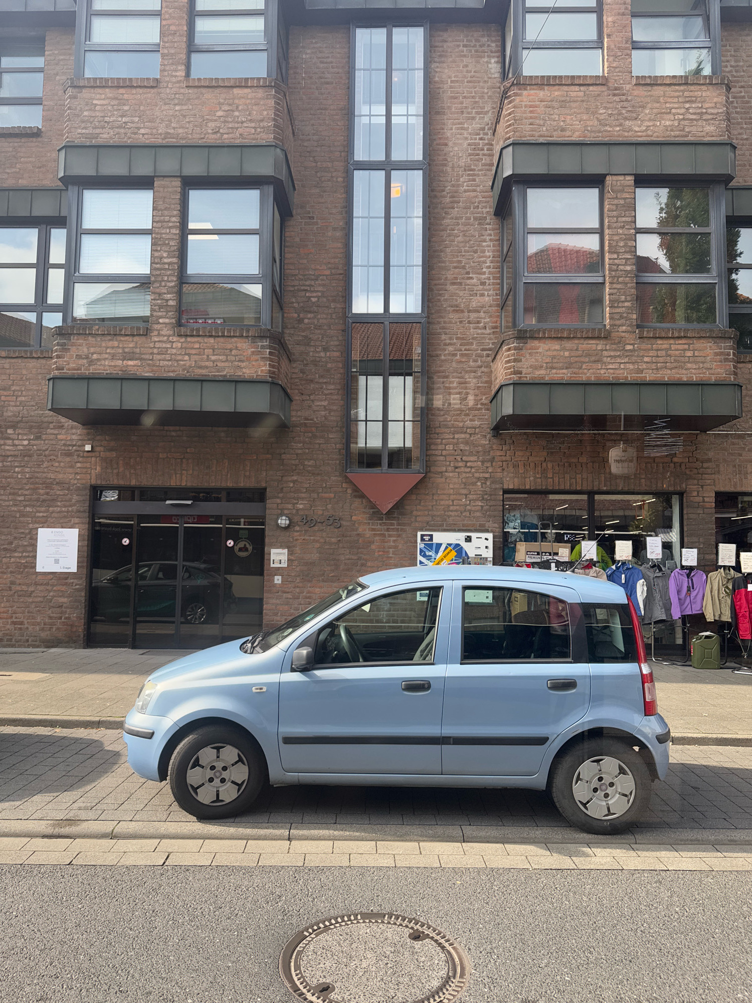 A light blue Fiat Panda is parked on the side of a street in front of a brick building in Cleves, Germany. The car is facing right, and its driver's side is visible. The building has multiple windows and a dark-colored entrance with automatic sliding doors. Clothes are displayed on racks outside a store on the ground floor of the building. The street in front of the building is paved with asphalt and cobblestones, and a circular manhole cover is visible in the foreground. The sky is overcast.