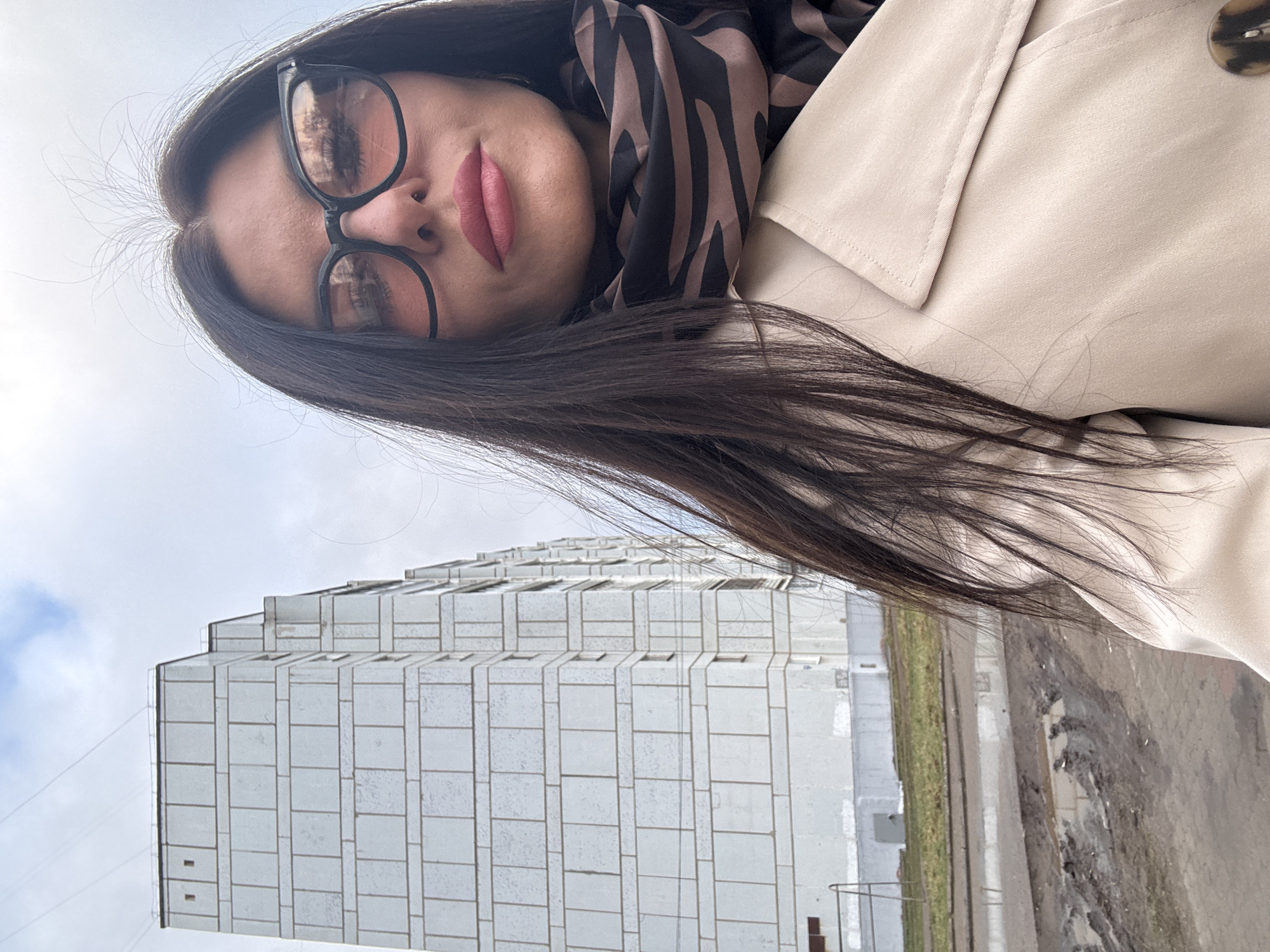 The image is a low-angle outdoor portrait, primarily featuring a woman in the foreground against an urban backdrop. The photograph is oriented 90 degrees counter-clockwise.
The subject is a woman with long, dark hair, which partially obscures her face. She wears dark-rimmed glasses and dark pink lipstick, with a neutral expression. She is dressed in a light beige trench coat or jacket, detailed with a large, mottled brown button on the lapel, and a black and brown striped scarf around her neck.
In the background, a large, light-colored (possibly white or light grey) multi-story building dominates the lower portion of the frame. The building features a grid-like facade with numerous rectangular panels and windows, indicative of utilitarian post-Soviet architecture, aligning with the provided context of Chaikovsky, Russia.
The ground in the foreground and mid-ground is a mix of dirt and muddy patches, along with dry, sparse grass or vegetation, suggesting damp conditions, possibly from recent rain. A concrete or paved edge runs alongside. The sky above is uniformly overcast and light grey, indicating a cloudy daytime setting. No visible text is present in the image.