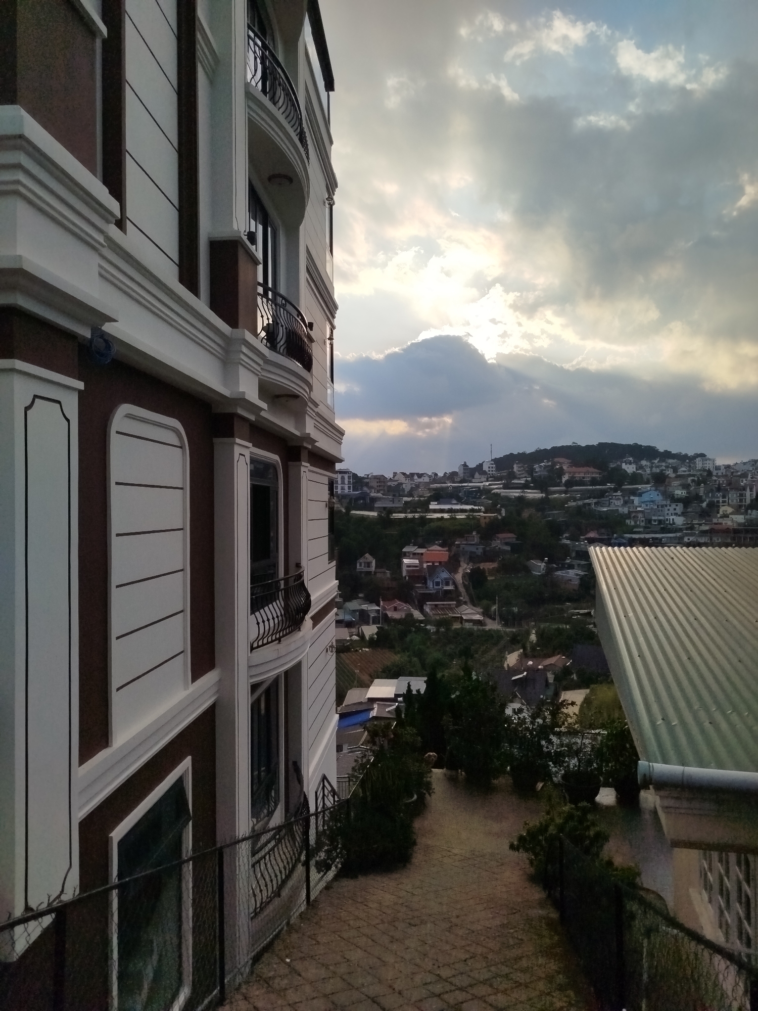 The image captures a view in Da Lat, Vietnam, characterized by a modern building in the foreground. The building features a brown and white facade with rounded balconies. The perspective is from an elevated position, looking down on a pathway and a roof with corrugated metal sheets. Beyond the pathway and roof, a cityscape unfolds across a valley with many small houses and buildings dotting the landscape. The sky is partly cloudy with rays of sunlight breaking through. No people are visible in the scene.