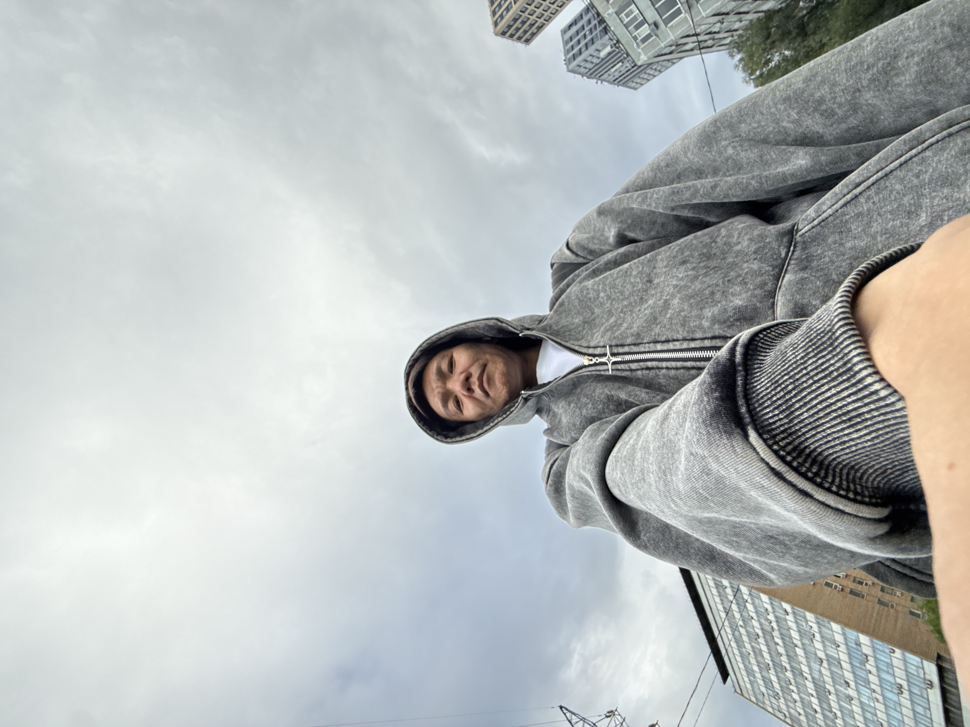 A person wearing a grey hooded jacket is taking a selfie from a low angle, looking up towards the cloudy sky. The background features several tall buildings, one a light brown brick building with many windows and air conditioning units, and another a modern grey skyscraper with a grid-like facade. There is also a large electrical pylon visible to the left of the frame. The weather appears overcast. The buildings suggest an urban environment, likely a city.