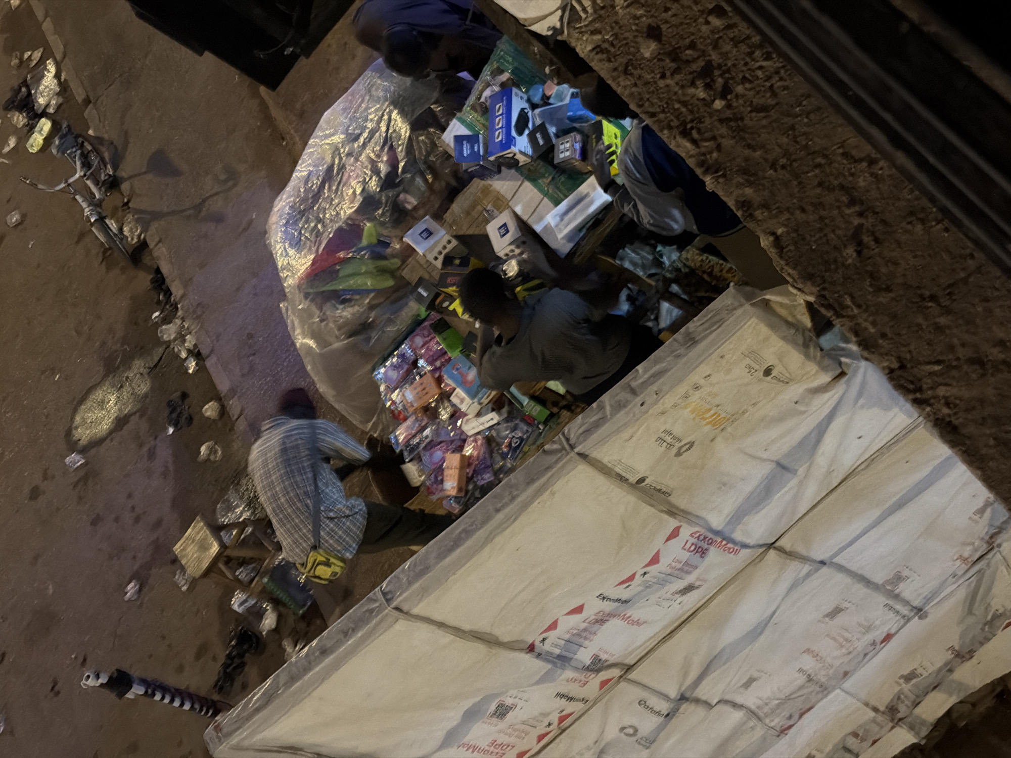 The image captures an outdoor scene at night, likely in Bukuru, Nigeria, with a focus on a street vendor's stall. Several individuals are present, appearing to be engaged in activities related to the stall. One person, wearing a checkered shirt and a yellow bag across their shoulder, is bent over, possibly attending to merchandise. Another person, in a grey shirt, is actively sifting through items at the stall. A third individual, in a blue and white top, is also near the stall, possibly a customer or assistant.
The stall itself is laden with various products, predominantly packaged electronics and small goods, displayed in baskets and boxes. A large, white, plastic-wrapped object, possibly a mattress or large packaging, dominates the foreground, partially obscuring the view of the street. The environment appears to be a street or market area, with a paved surface showing signs of wear and scattered debris. In the background, a bicycle lies on its side, and a street light with a distinctive black and white striped pole is visible. The overall impression is of a busy, informal commercial setting at night.