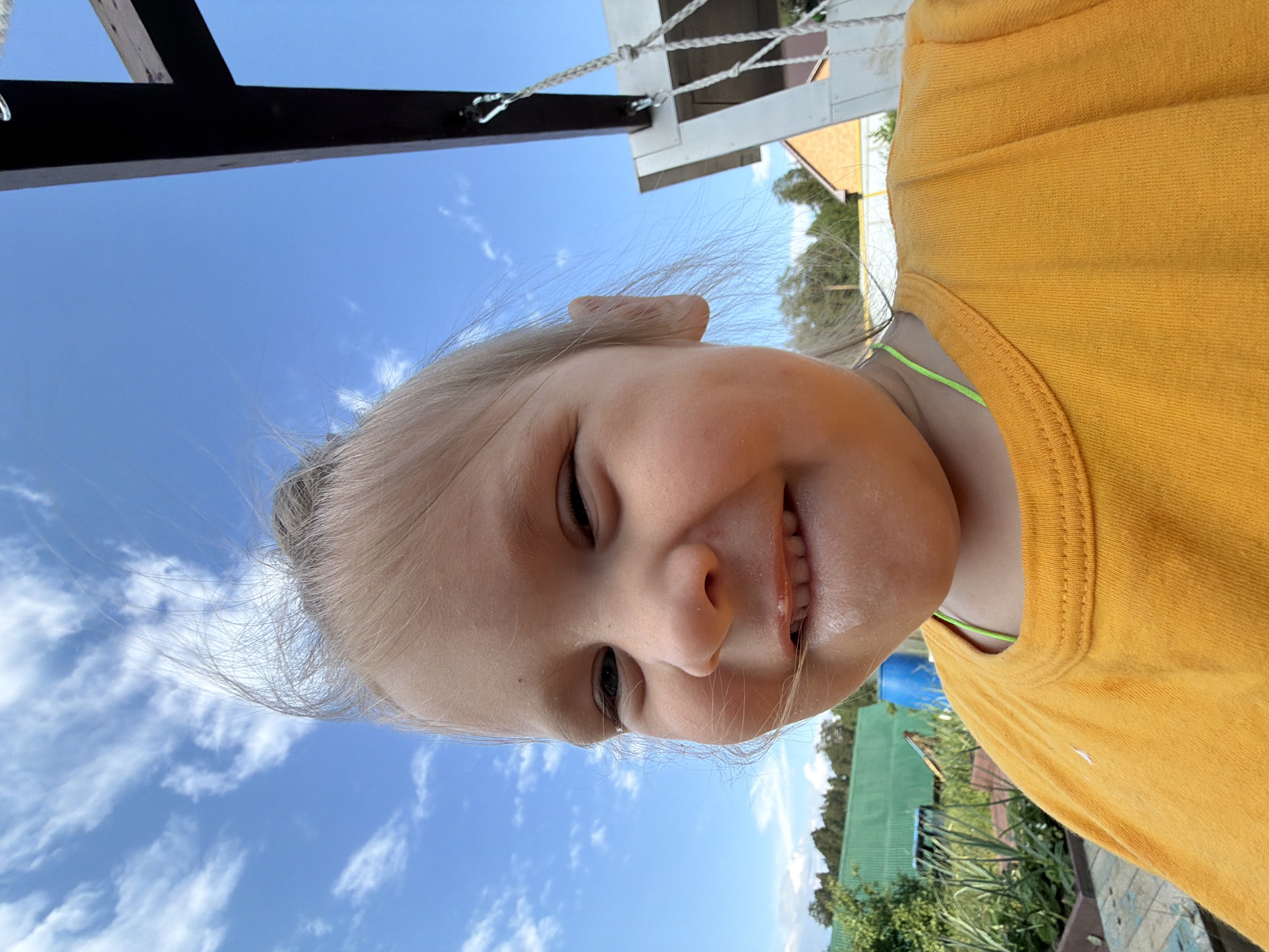 A close-up, low-angle shot captures a young child with fair hair pulled up, smiling broadly under a bright blue sky with scattered white clouds. The child wears an orange t-shirt. The background shows a green wooden structure, possibly part of a playground or outdoor seating area, and a hint of a residential building with an orange roof. The scene appears to be outdoors in a rural or suburban setting, likely on a sunny day. The child's expression suggests happiness and playfulness. The overall impression is one of casual, outdoor recreation.