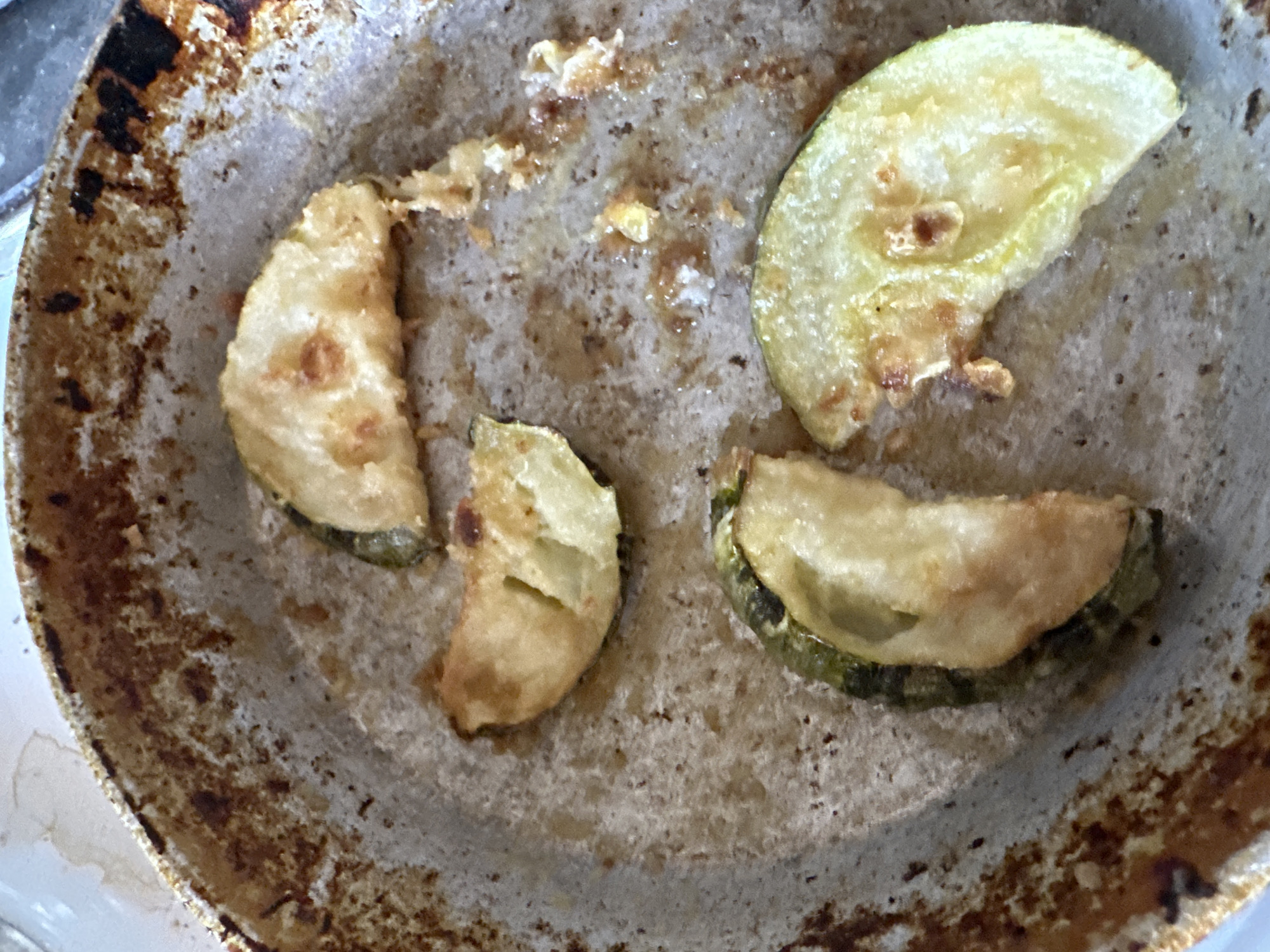 The image shows slices of fried zucchini in a dirty, used frying pan. The pan is made of a metallic material and has a seasoned, non-stick surface that is heavily stained and shows signs of wear and tear, with dark brown and reddish-brown burnt residues. Four slices of zucchini are visible, each appearing partially cooked or fried to a golden-brown color. The zucchini slices are cut into rounds, and some have a green rind. The pan is situated on a light-colored, textured surface, possibly a countertop or table. There are no people or other objects present in the image, and the scene appears to be indoors, likely in a kitchen. The location of Biysk, Russia is not evident from the visual information provided in the image.