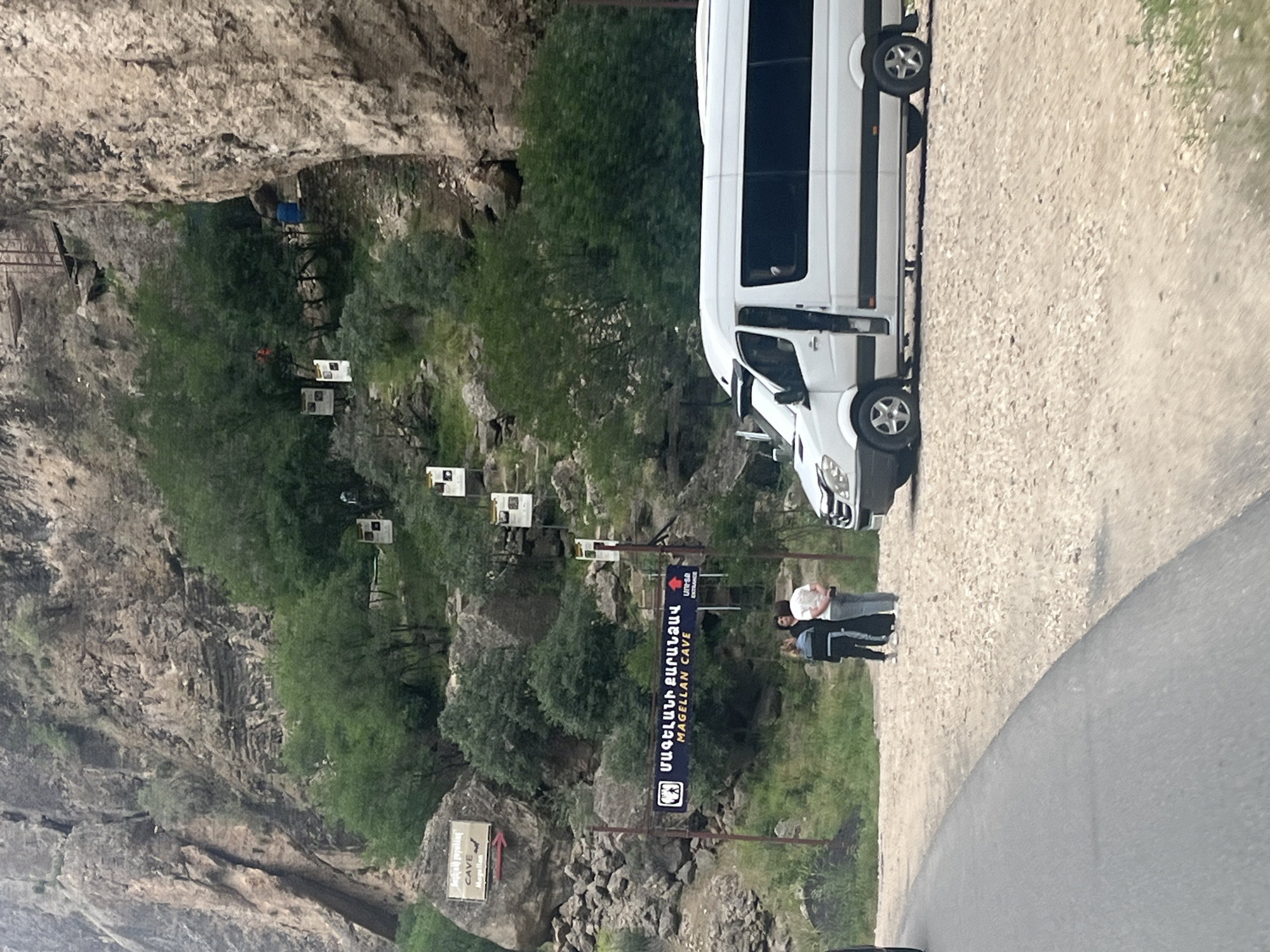 The image captures the entrance to Magellan Cave in Areni, Armenia, with a significant rocky and vegetated mountainside forming the backdrop. A large dark blue sign with white text, reading "ՄԱԳԵԼԼԱՆԻ ՔԱՐԱՆՁԱՎ" and "MAGELLAN CAVE," is centrally positioned, indicating the attraction. Below this sign, three individuals are standing on a light-colored gravel surface, seemingly engaged in conversation. One person on the left wears a dark jacket and pants, the middle person is in a dark long garment, and the person on the right wears a light top and light pants.
To the right of the individuals, a white Mercedes-Benz Sprinter van is parked on the gravel, with its passenger side door open. Another white vehicle is partially visible behind it. On the far left, a segment of a dark vehicle is visible on a paved road that curves into the frame.
Further up the mountainside, a smaller rectangular sign on a large rock reads "CAVE Magellan" with an arrow pointing right and a bat silhouette. Multiple smaller white informational signs are sporadically placed along a winding path that ascends the vegetated slope. A wooden structure, possibly stairs or a platform, is visible high on the cliff face to the right. The terrain is rugged, characterized by large rock formations and a mix of green trees and shrubs. The lighting suggests an overcast sky.