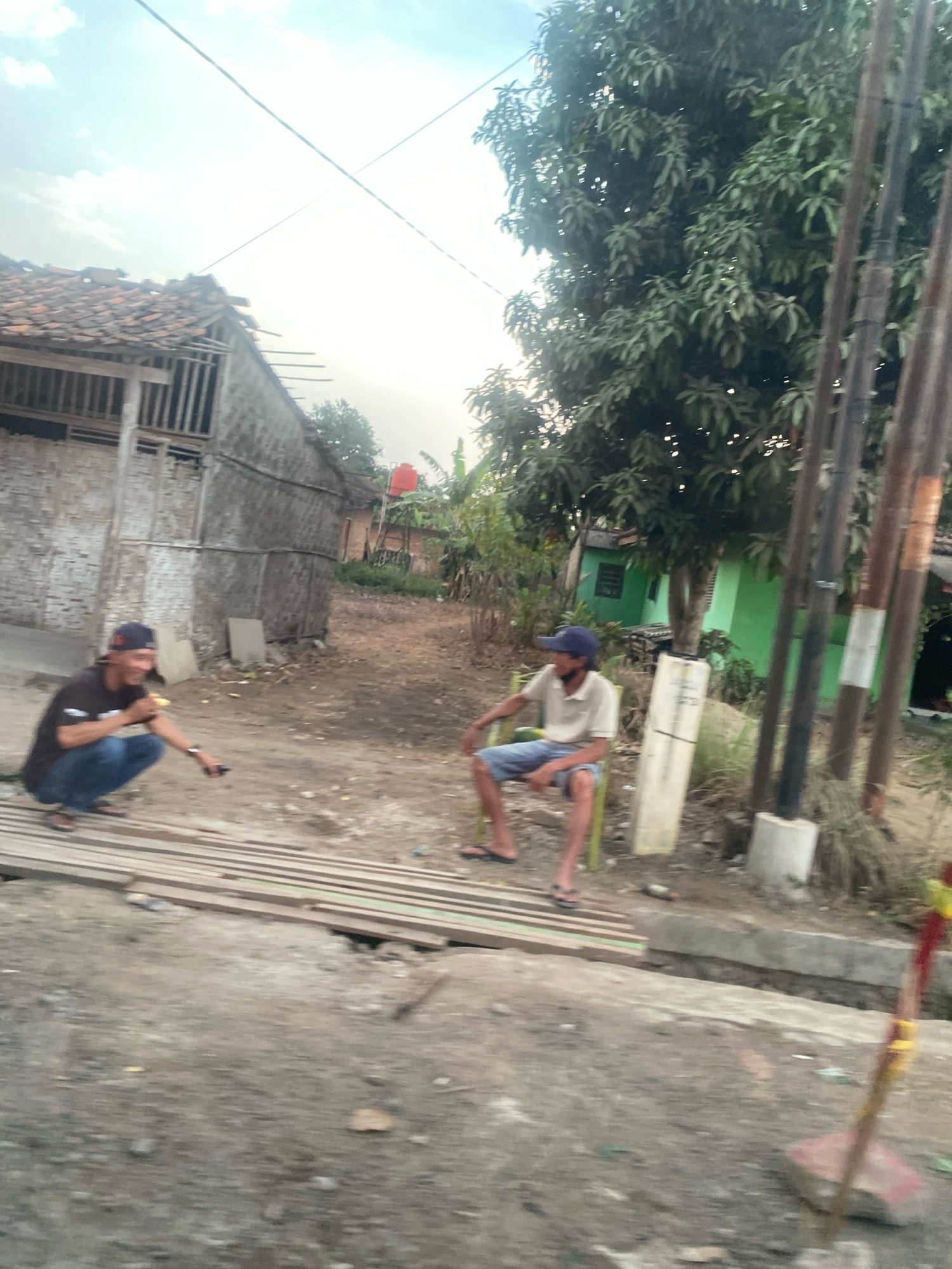 In Banjar Sari, Indonesia, two individuals are observed in a rustic outdoor setting. The person on the left, wearing a black t-shirt and blue jeans, is crouched down, holding a yellow object and pointing with their right hand towards the ground. They have a dark baseball cap on. To their right, a second person is seated on a green chair, wearing a light-colored polo shirt and blue shorts, and a dark baseball cap. They appear to be looking towards the left. In the background, a dilapidated building with a tiled roof is visible, alongside a green structure and lush trees. Several utility poles with wires are also present. The ground is covered in dirt and rubble, with some wooden planks laid out in the foreground. The overall scene suggests a casual outdoor activity in a somewhat undeveloped area.
