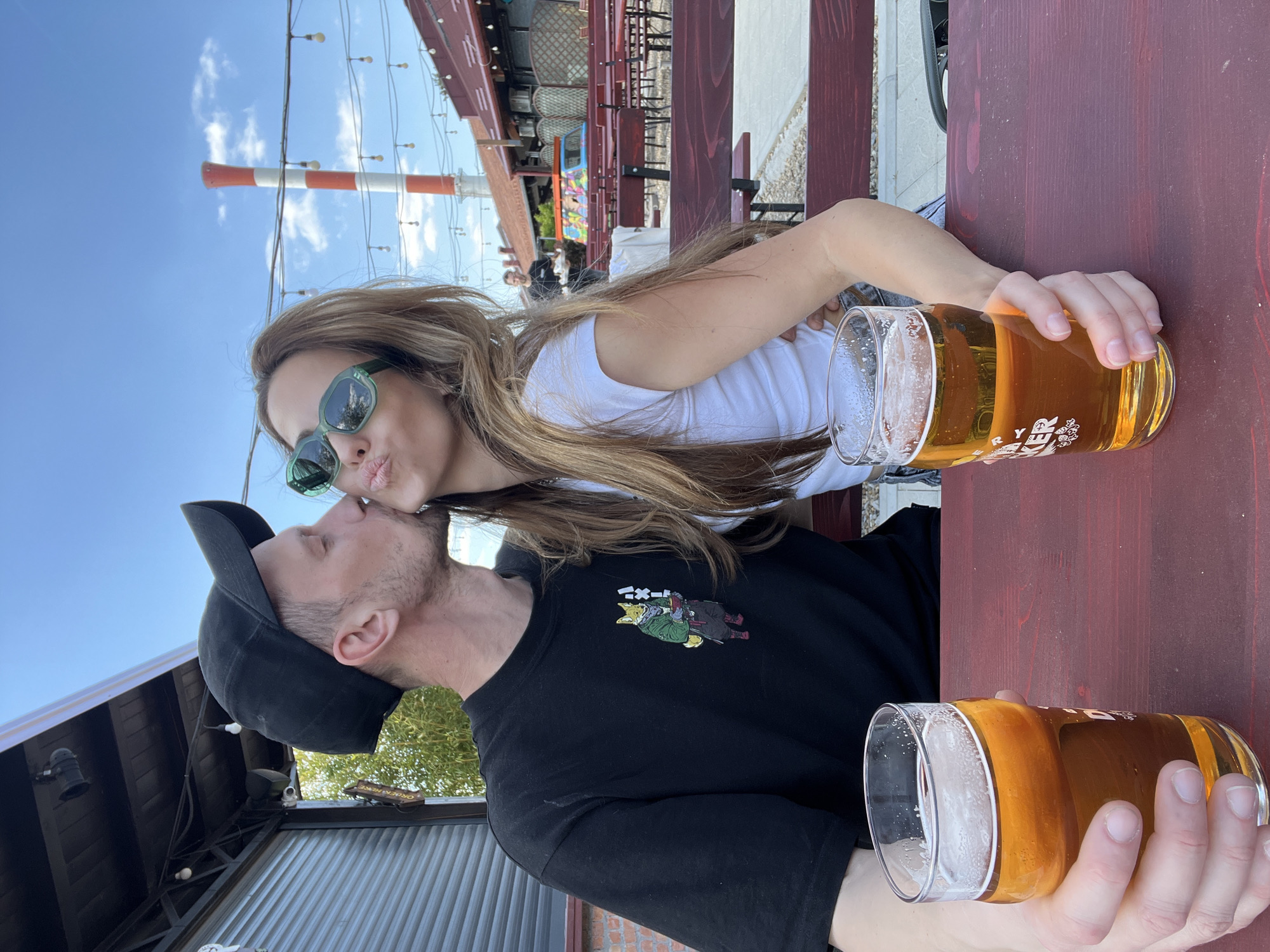 A photograph depicts a couple at an outdoor seating area of a restaurant or bar in Belgrade, Serbia. The woman, with long brown hair and wearing green sunglasses and a white t-shirt, is leaning over to kiss the man, who is wearing a black t-shirt and a baseball cap. They are seated at a wooden table. Each of them is holding a full glass of beer. Behind them, there are tables and other people in the outdoor area. In the background, a red-and-white striped industrial chimney is visible against a blue sky.