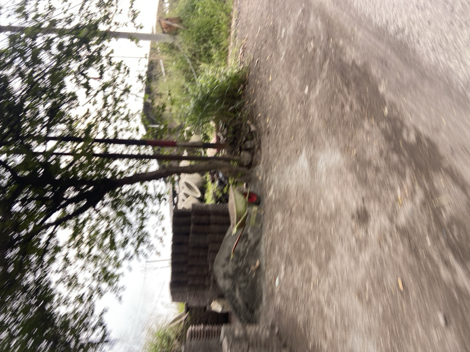 The image shows a dirt road in Kecamatan Margaasih, Indonesia, at the coordinates -6.961 latitude and 107.536 longitude. A large tree is in the foreground, with a pile of gravel and a wheelbarrow to the left. Behind them is a stack of cinder blocks and a few other large, circular objects. The image is taken from a moving vehicle, giving it a blurry appearance. The road continues into the distance, where there are trees and foliage on both sides. There are no people visible in the image.