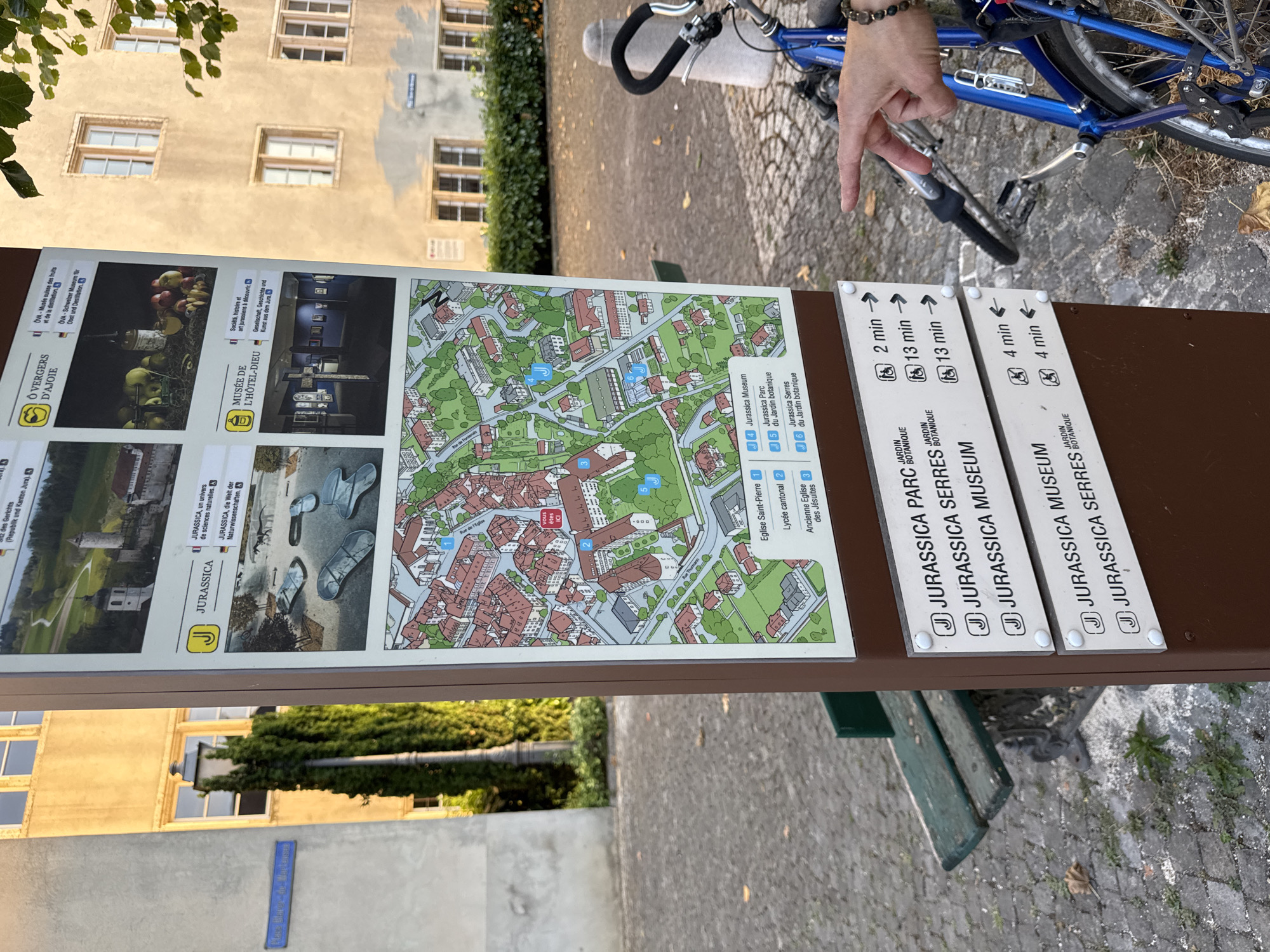 The image shows a detailed information signpost in Porrentruy, Switzerland. The signpost is positioned outdoors in a paved area, with a park bench and lush green trees visible in the background. To the right of the signpost, a blue bicycle with a dark handlebar is parked, and a person's hand with a bracelet is pointing towards the sign. The signpost features a large map of the city center with various points of interest marked with numbers and letters. Below the map, there are lists of attractions, including "JURASSICA MUSEUM" and "JURASSICA PARC du Jardin Botanique," with estimated walking times indicated. The architecture of the surrounding buildings, visible in the background, suggests a historic European city. The lighting indicates it is daytime, with sunlight casting shadows and highlighting the textures of the paving stones and foliage.