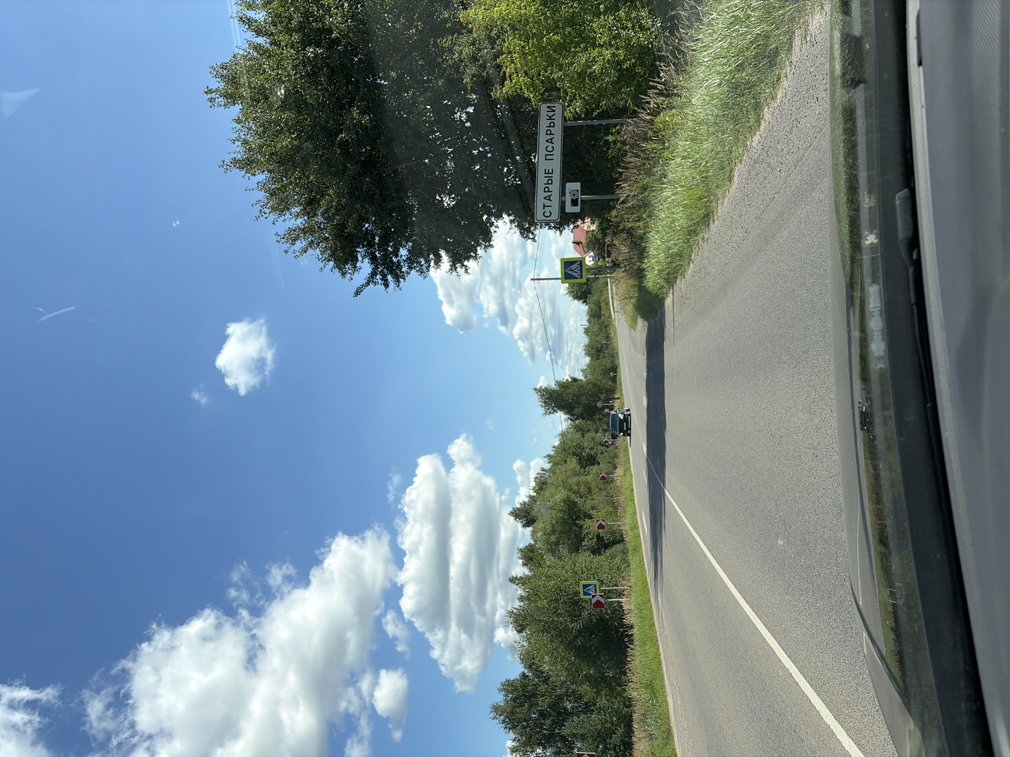 The image depicts a view from inside a vehicle driving on a paved road in Starye Psar'ki, Russia. The road curves to the left, with a white line marking the lane division. On the right side of the road, a sign reads "СТАРЫЕ ПСАРЬКИ," indicating the village name, and below it, a camera symbol suggests a speed camera. Adjacent to this is a pedestrian crossing sign. Lush green trees and grass line the roadside. Several red and white directional signs are visible along the road, indicating curves. A black car is driving ahead on the road. The sky is a bright blue with scattered white clouds. The foreground shows the car's dashboard and windshield.