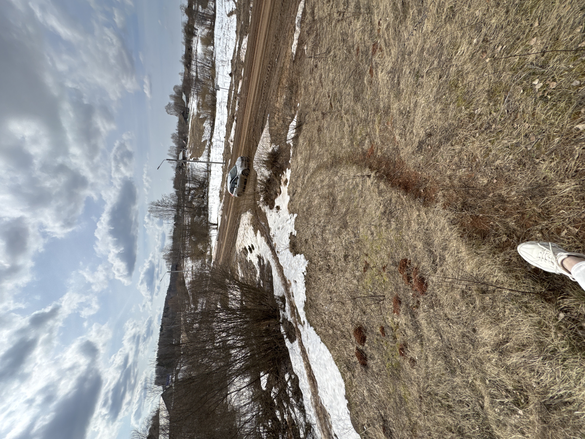 The image shows a rural scene, potentially in Vassyata, Russia, given the prompt's information. A dirt road runs through the landscape with patches of snow visible on either side. A light-colored vehicle is parked near the road. The terrain transitions into a field of dry grass. Bare trees and bushes are present, and a section of the sky with clouds is visible. A person's white shoe is visible on the right side of the image, suggesting the photo was taken from a low angle.