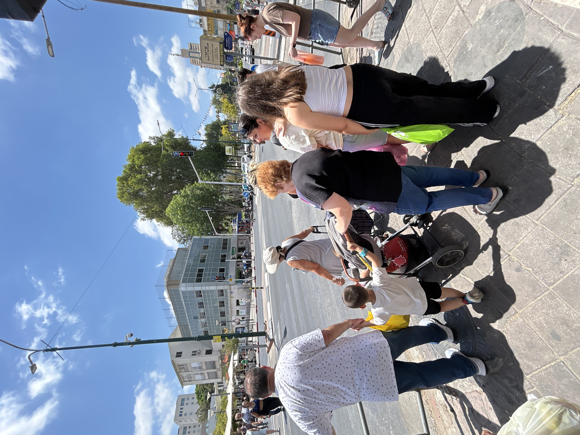 In Rishon LeZion, Israel, on a clear day with scattered clouds, a group of people is captured on a sidewalk next to a street. The foreground features a man in a white patterned shirt and blue jeans holding hands with a young boy in a white t-shirt and black shorts. Behind them, a woman in a wide-brimmed hat and a grey sleeveless dress walks with a stroller, holding its handle. To the right, a woman in a white tank top and black athletic pants stands with a bright green shopping bag. Another woman, wearing a brown t-shirt and denim shorts, walks past in the background. Several other individuals and pedestrians are visible further down the street and on the opposite sidewalk. Buildings, trees, traffic lights, and street lamps form the urban environment. The image seems to be taken during daylight hours, judging by the bright sunlight and the distinct shadows cast on the paved ground.