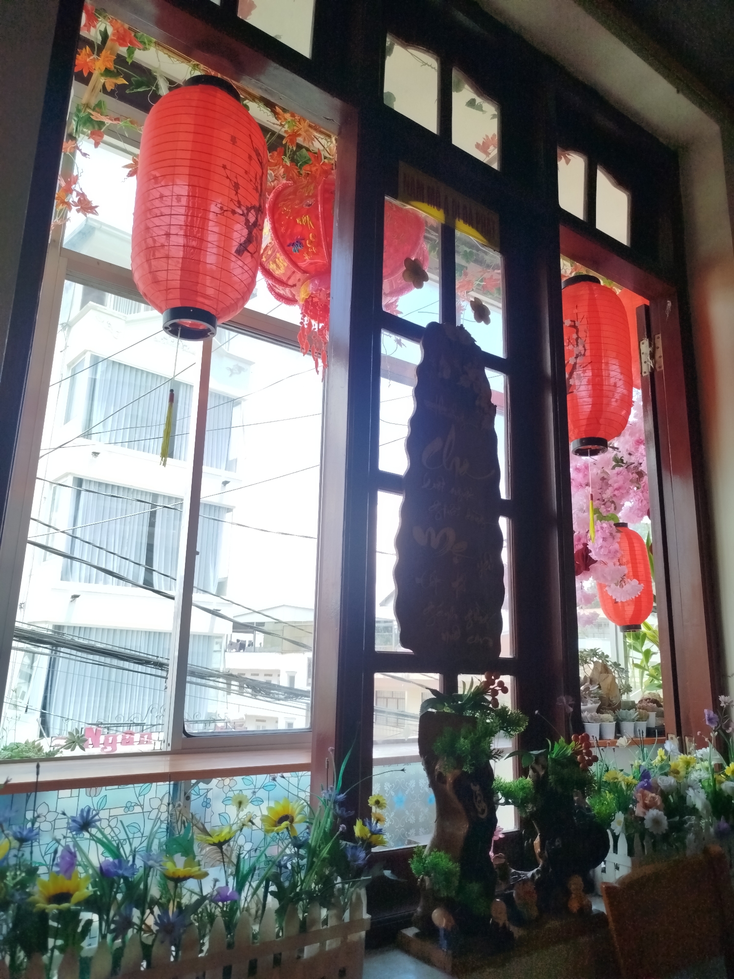 A medium shot captures a brightly lit indoor scene, looking out through a decorative window towards an urban landscape. The interior is adorned with cultural elements, notably two red lanterns hanging in the window. The window itself is divided into sections, some of which are open, revealing the exterior. A wooden sign with inscribed text in Vietnamese is visible in one section. Below the window, a display of artificial flowers in a white picket fence planter adds color to the scene, with two decorative, miniature sculpted tree root sculptures in front of it.
Outside, the view encompasses a white building with multiple windows and utility lines crisscrossing the sky. The daylight suggests it is daytime. The scene appears to be located in Da Lat, Vietnam, due to the architectural style and decorative elements commonly found in the region. There are no visible people or interactions captured in the frame.