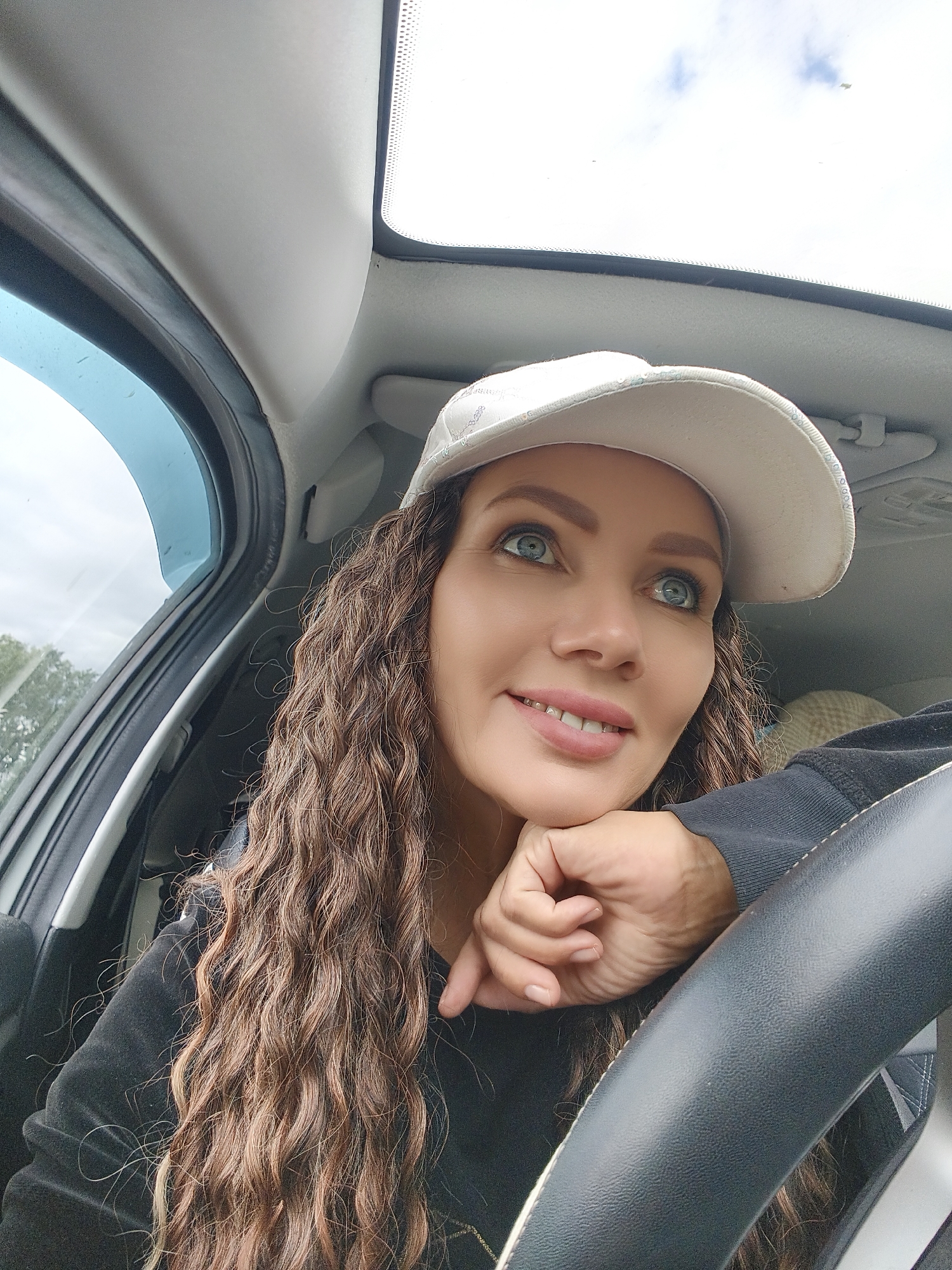 A medium shot captures a woman with long, curly brown hair and blue eyes wearing a white baseball cap and a black sweatshirt. She is leaning her head on her hand, which is resting on a car's steering wheel, and looking upwards and to the right. Her mouth is slightly open in a smile, and her eyes are wide. The interior of the car is visible, including the roof with a sunroof, the car door with a blue tint, and the steering wheel. The background outside the car appears to be overcast with some trees visible. The lighting is bright, suggesting it is daytime.