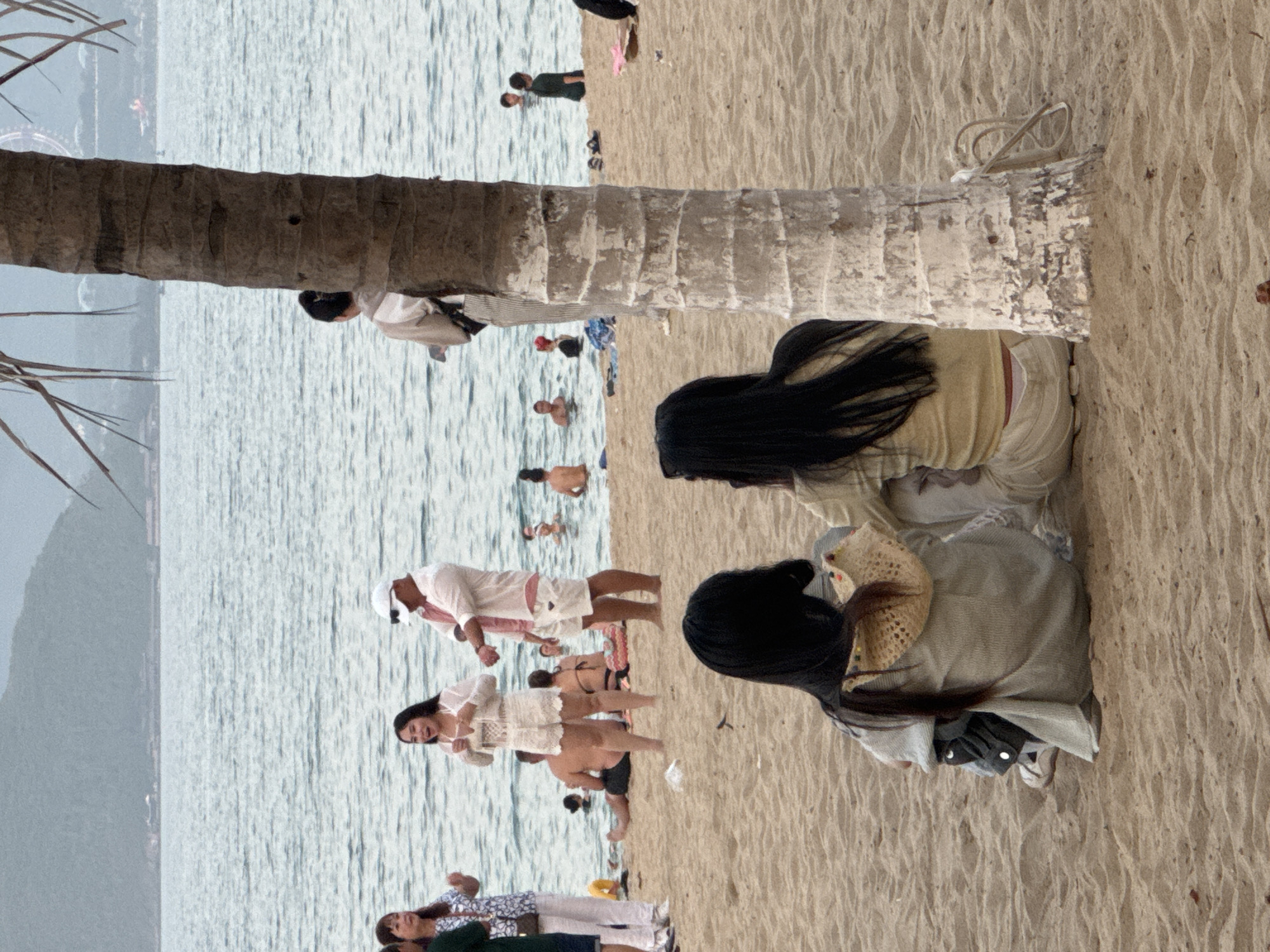 A wide shot captures a sandy beach scene in Nha Trang, Vietnam, with the ocean and a distant hazy coastline in the background. In the foreground, two women with long black hair are seated on the sand, facing away from the camera, with their backs to a large palm tree trunk. The woman on the left wears a light-colored jacket over a darker garment and has a woven hat on her shoulder. The woman on the right wears a light-colored top and pants.
In the middle ground, several people are present on the beach and in the shallow water. A man in a white shirt and shorts, wearing a white cap, stands near the water's edge. A woman in a white outfit, also near the water, gestures with her hands and appears to be talking. Other individuals are scattered on the sand and swimming or standing in the ocean. A palm tree trunk stands prominently in the right side of the frame, partially obscuring some of the middle-ground elements. The water is calm and reflects the overcast sky. The sand is a light tan color with visible footprints and some scattered debris.