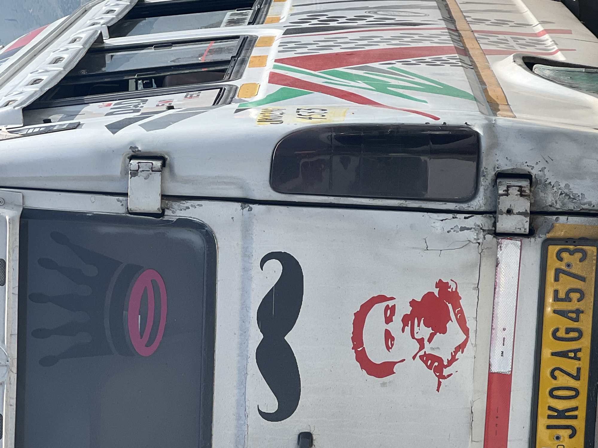 The image shows the rear and side of a white bus, likely in Ujroo, India, as indicated by the license plate "JK02AG4573". The bus has several decals. On the rear window, there is a dark gray silhouette of a crown with a pink oval shape within its band. Below the window, a black silhouette of a handlebar mustache is centered. Beneath the mustache, a red stencil of a man's face is depicted, with him appearing to hold a cigarette. A reflective red and white strip is visible above the license plate. The side of the bus features a series of colorful diagonal stripes in red, green, and white, along with some text that is partially obscured but appears to include "JK02AG 4573". The bus shows signs of wear, with peeling paint and rust on the hinges. The background suggests an outdoor setting, with a hint of mountains in the distance.