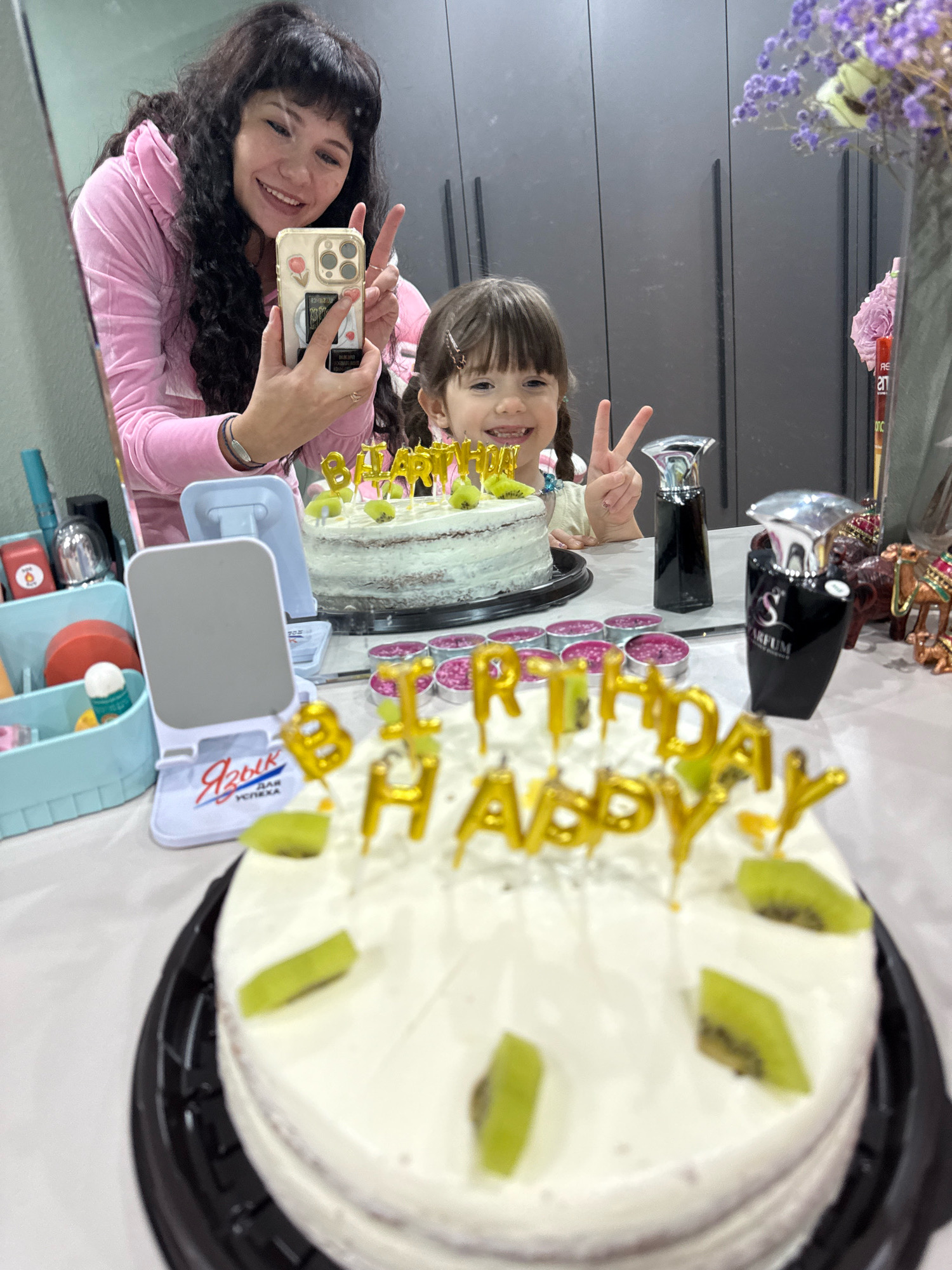 A woman in a pink velour tracksuit is taking a selfie in a mirror, holding her phone with a sticker-adorned case. Next to her, reflected in the mirror, is a young girl with braided hair, smiling and holding up two fingers in a peace sign. In the foreground, two cakes are visible. The larger cake, in sharp focus, is white with gold "HAPPY BIRTHDAY" candles and slices of kiwi. A smaller cake, slightly out of focus, is behind the girl and also appears to have birthday candles. Various items are arranged on a table, including small pink tins, perfume bottles, decorative camels, and a makeup organizer. The background shows a mirrored closet and a vase with purple flowers. The scene suggests a birthday celebration, possibly in a home environment. The location, Cheboksary, Russia, is implied by the presence of a white stand with Cyrillic text that translates to "Language of Success".
