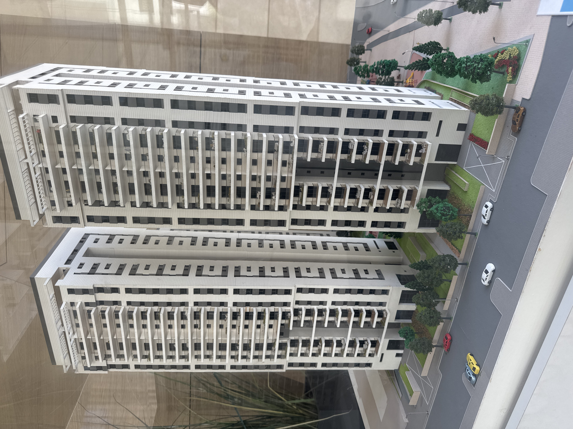 The image shows an architectural model of a modern building complex. The buildings appear to be high-rise residential structures with a repeating pattern of windows and balconies. The model includes landscaping details such as trees, grass, and miniature cars on a street. The model is viewed through glass. The style and scale of the model suggest it represents a future development or a conceptual design. The details suggest the design could be for a residential development or apartment complex. The location is possibly in Netanya, Israel.