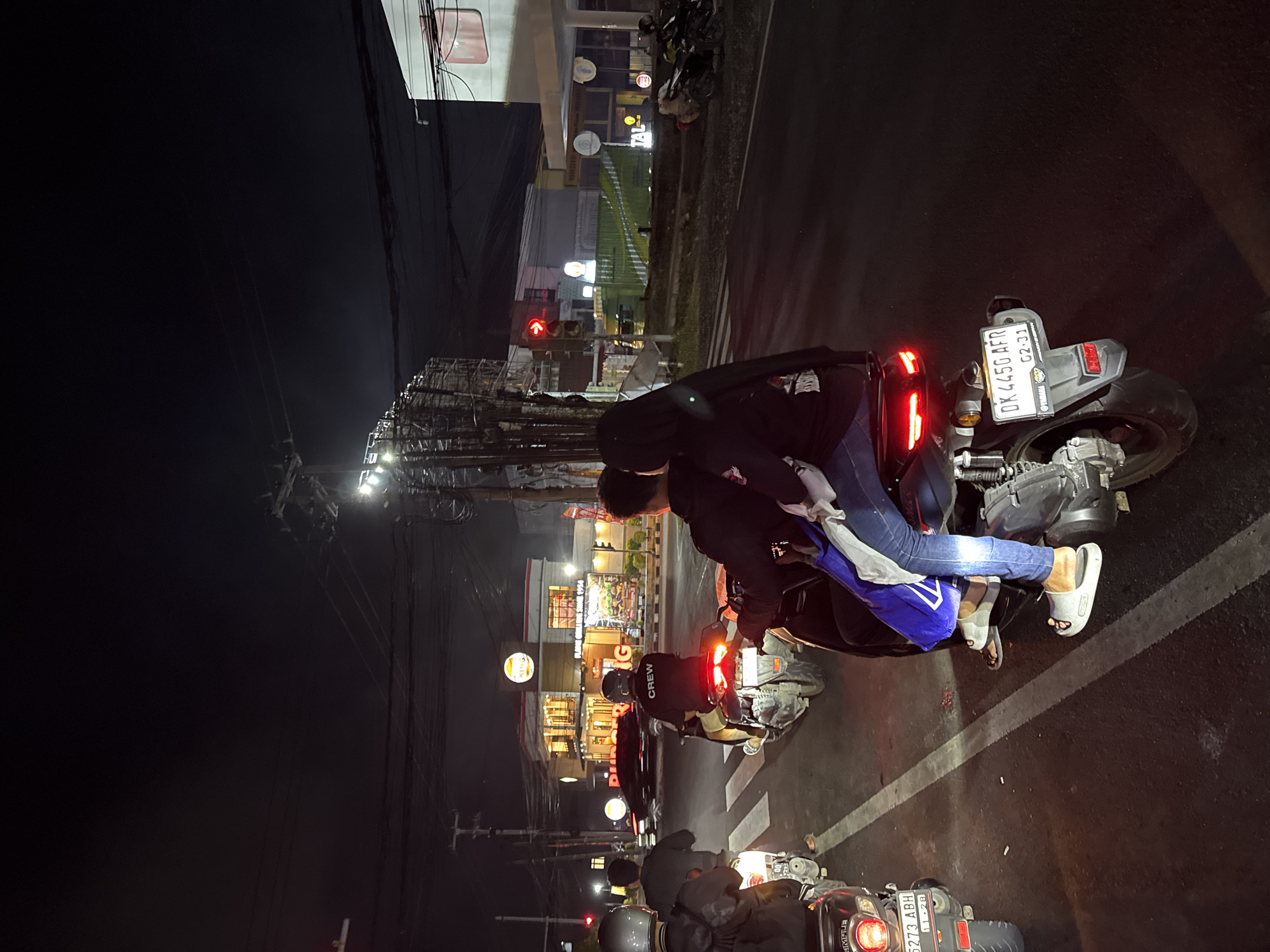 This image depicts a nighttime scene on a city street in Denpasar, Indonesia. The foreground is dominated by several motorcycles, with two in sharp focus. On the right, a motorcycle is ridden by a person wearing blue jeans and white sandals, with another person behind them wearing a black hijab and a black jacket. This motorcycle has a license plate that reads "DK 4450 AFR" and the registration date "02-21". To the left, another motorcycle is visible, its rear red taillight illuminated. Further down the road, more motorcycles and their riders are present, some with helmets.
In the background, a Burger King restaurant is prominently lit, with its iconic signage visible. The street appears to be a multi-lane road with traffic lights and overhead electrical wires. Buildings line the side of the street, some with illuminated windows. The overall atmosphere is that of a busy urban environment at night.