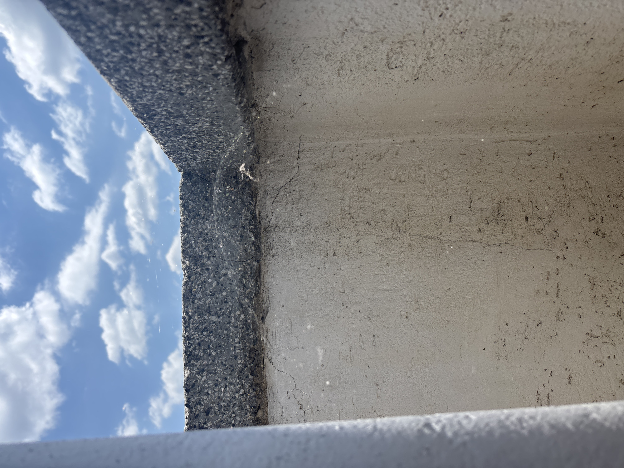 The image displays a close-up view of a textured, light-colored concrete wall with a darker, rough-textured concrete coping at the top. The scene is outdoors, with a bright blue sky and scattered white clouds visible in the background. A white cylindrical object, possibly a pipe or railing, is partially visible on the left side of the frame. The concrete wall exhibits some minor cracking and discoloration, suggesting age or weathering. A small spider web with a spider is visible on the top edge of the wall, near the right corner of the frame. The composition is focused on the architectural elements and the sky, with no people or other prominent objects in view. The location is identified as Zlatograd, Bulgaria.