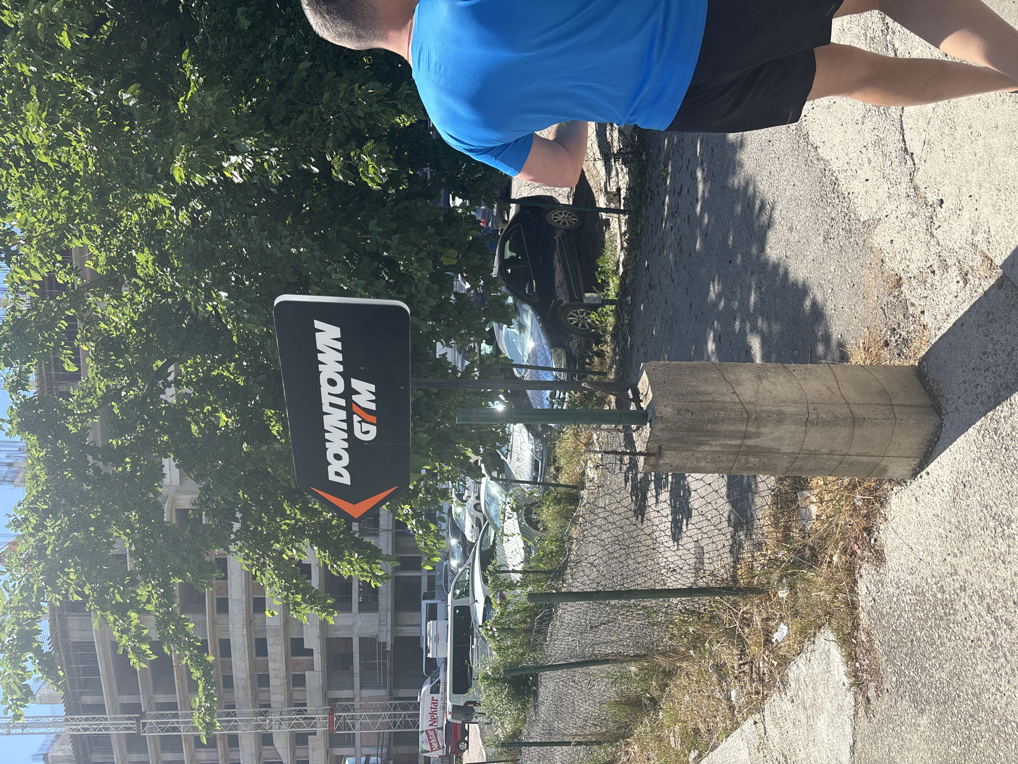 A medium shot captures a man wearing a blue shirt and black shorts walking along a sidewalk beside a "Downtown Gym" sign in Budva, Montenegro. The sign is black with an orange accent and is erected next to a concrete base and a chain-link fence. Behind the fence, several parked cars can be seen. In the background, there are trees and an unfinished high-rise building under construction. The sidewalk is paved with concrete and shows signs of wear. Sunlight casts shadows across the scene.