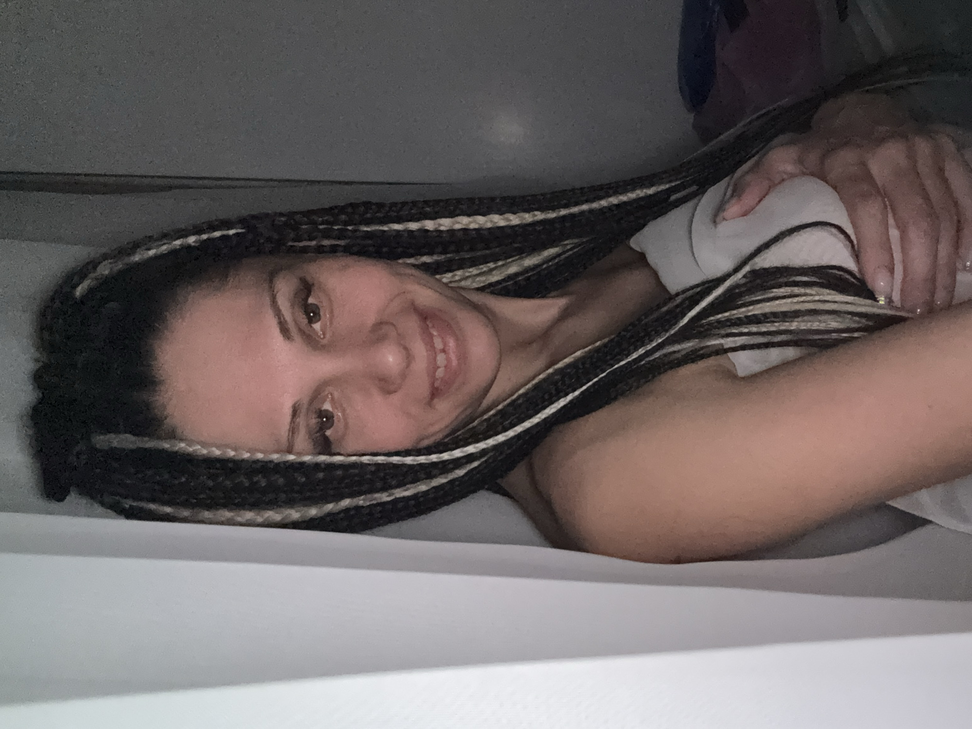 The image displays a close-up, portrait-oriented shot of a person, likely a woman, lying down. The perspective is from above, looking down at the individual who is smiling up at the camera.
**Subjects:**
The primary subject is a person with long, dark hair styled in braids, featuring prominent lighter (possibly white or blonde) highlights interwoven throughout. Their face is turned towards the camera, revealing a broad smile. One arm is visible, resting on a light-colored blanket or sheet, with the hand partially clasped around it. The background is dark and indistinct, suggesting an indoor setting with low lighting.
**Activity or event taking place:**
The person is lying down, presumably on a bed or a soft surface, and appears to be posing for the camera. Their smile conveys a sense of happiness or contentment.
**Notable details:**
- **Hair:** The most prominent feature is the intricate braided hairstyle, combining dark and light strands.
- **Face:** The person has expressive dark brown eyes, visible eyelashes (possibly enhanced with mascara), and a bright, open smile.
- **Hands/Nails:** The visible hand shows fingers with manicured nails, painted with a light, possibly glittery, polish.
- **Lighting:** The overall lighting is dim, indicating it might be evening or night, or a room with subdued illumination.
- **Attire/Bedding:** A light-colored, possibly white, sheet or blanket covers parts of the body.
- **Emotion:** The clear smile suggests a positive and relaxed mood.
- **Location Cues:** There are no specific visual cues within the image itself to suggest the location is Rusanovo, Russia, as provided in the context. The setting appears to be a generic indoor space.
- **Visible Text:** No visible text is present in the image.
The scene captures a moment of casual relaxation and happiness.