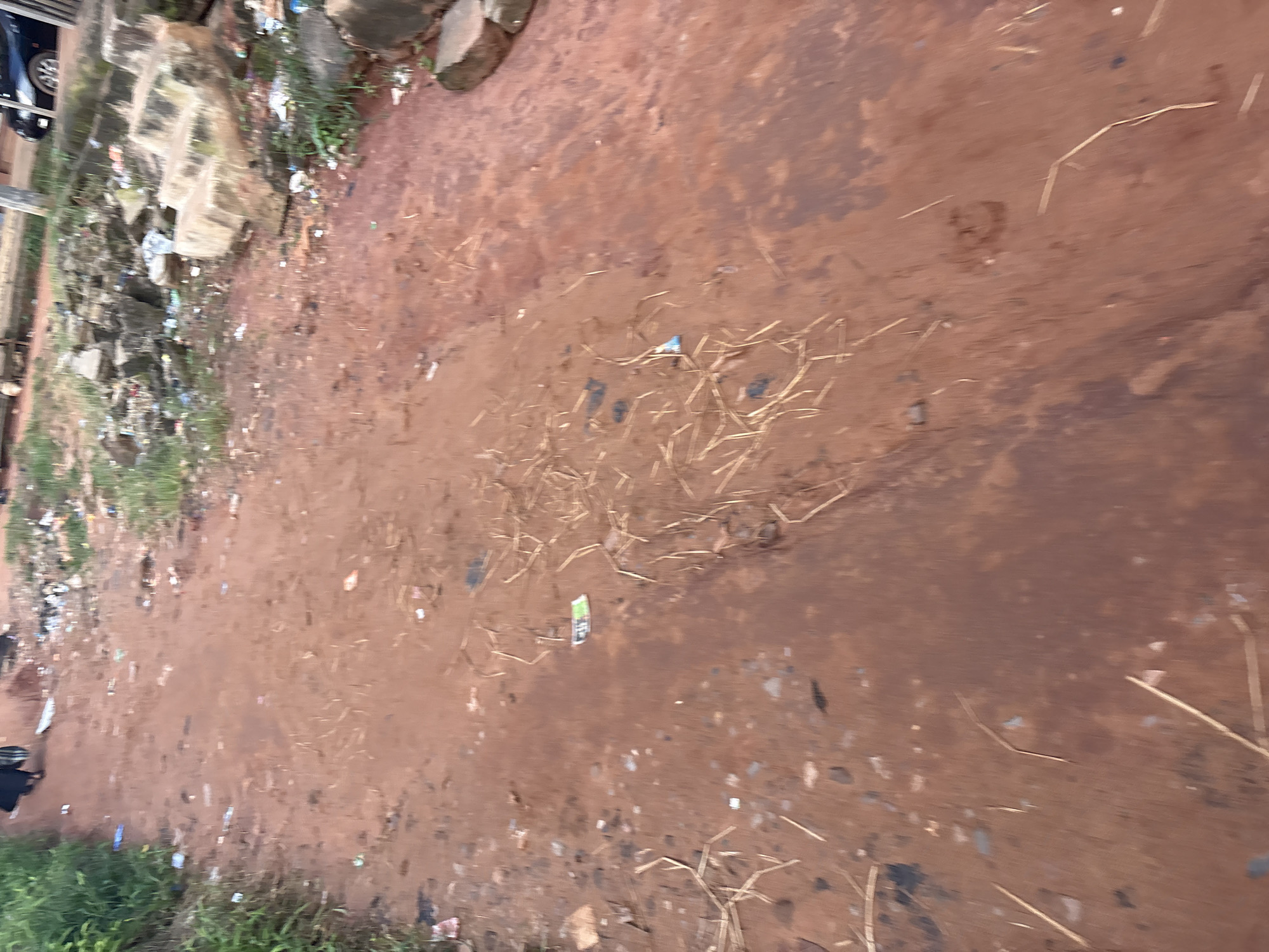 The image depicts a narrow, unpaved path made of reddish-brown earth, likely in Onitsha, Nigeria, as indicated by context. The path is littered with various debris, including dry grass, small stones, and scattered pieces of trash such as plastic wrappers and discarded paper. To the left of the path, there is a dense growth of green vegetation. In the background, on the right side, there are piles of stones, possibly part of a construction or a natural rock formation, also with visible trash. Further in the background, a dark-colored vehicle is parked, and there are indications of buildings or structures. A person wearing dark clothing, possibly a long skirt and shirt, is partially visible walking away from the camera on the upper left side of the path. The lighting suggests it is daytime, possibly overcast or late afternoon given the diffused light.