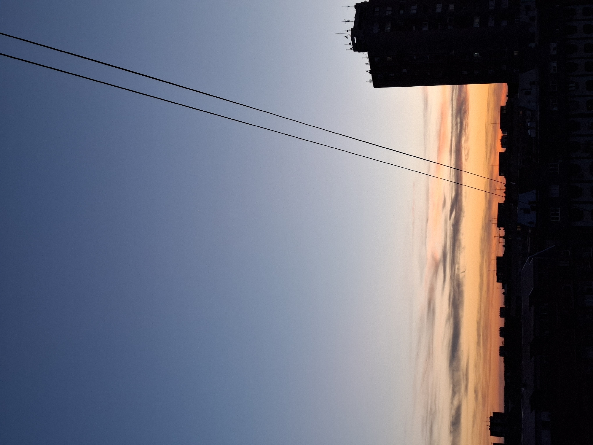 The image shows a view of the sky during sunset, framed by urban elements. The sky transitions from deep blue on the left to orange and pink hues where the sun sets. A building silhouette is visible on the right side. Two parallel cables stretch across the upper left portion of the sky. The location is Novi Sad, Serbia.