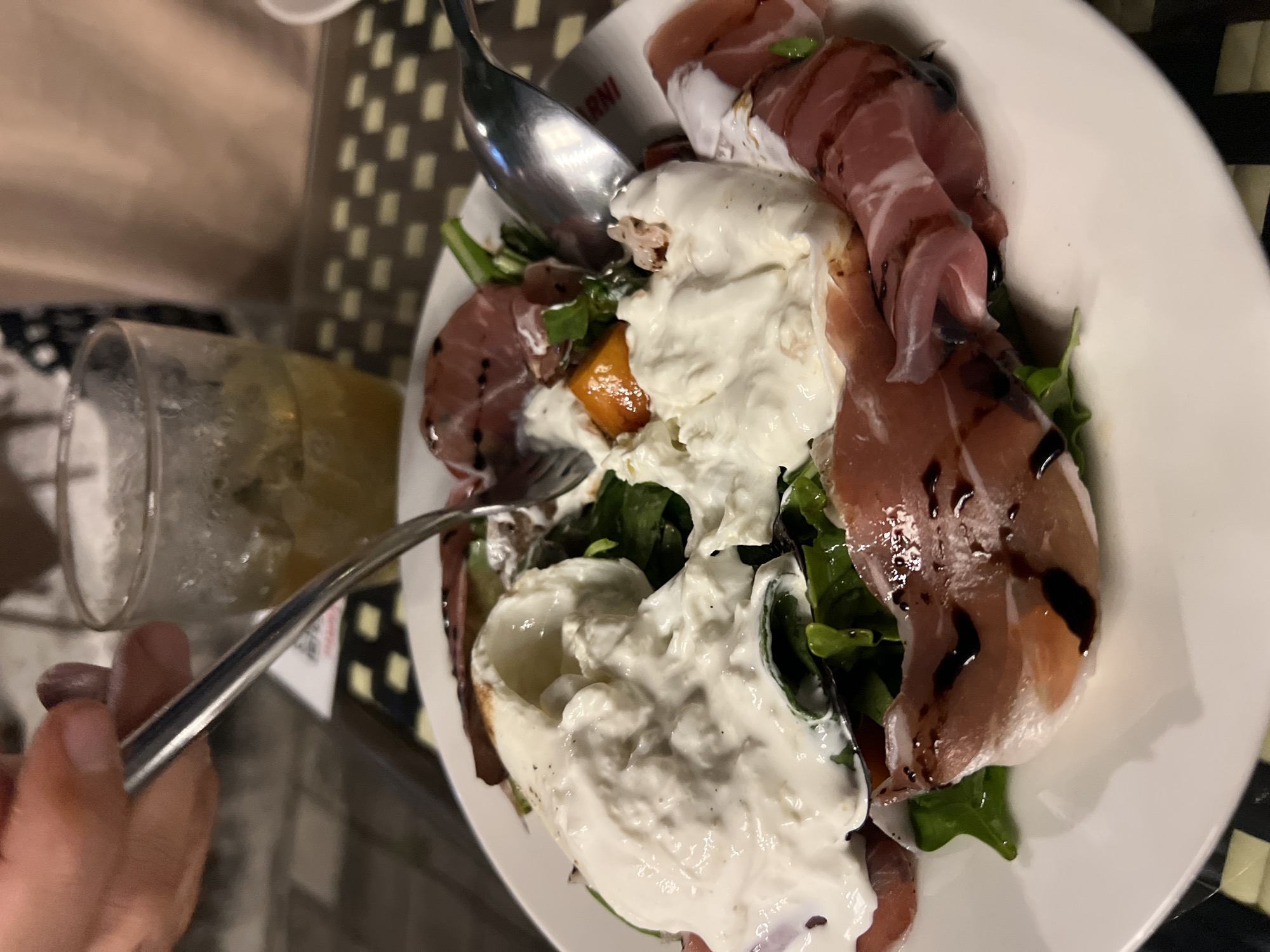 A top-down, close-up shot depicts a person eating a salad in what appears to be a restaurant setting. The salad is presented on a white plate and features prosciutto, leafy greens, a pale creamy cheese, and an orange item. The salad is dressed with a dark balsamic glaze. A glass containing a yellow liquid is visible on the left. A patterned surface that could be a tabletop or placemat is present. An individual's hand is holding a fork and a spoon is also inside the plate. The location in the city of Unknown, Unknown.