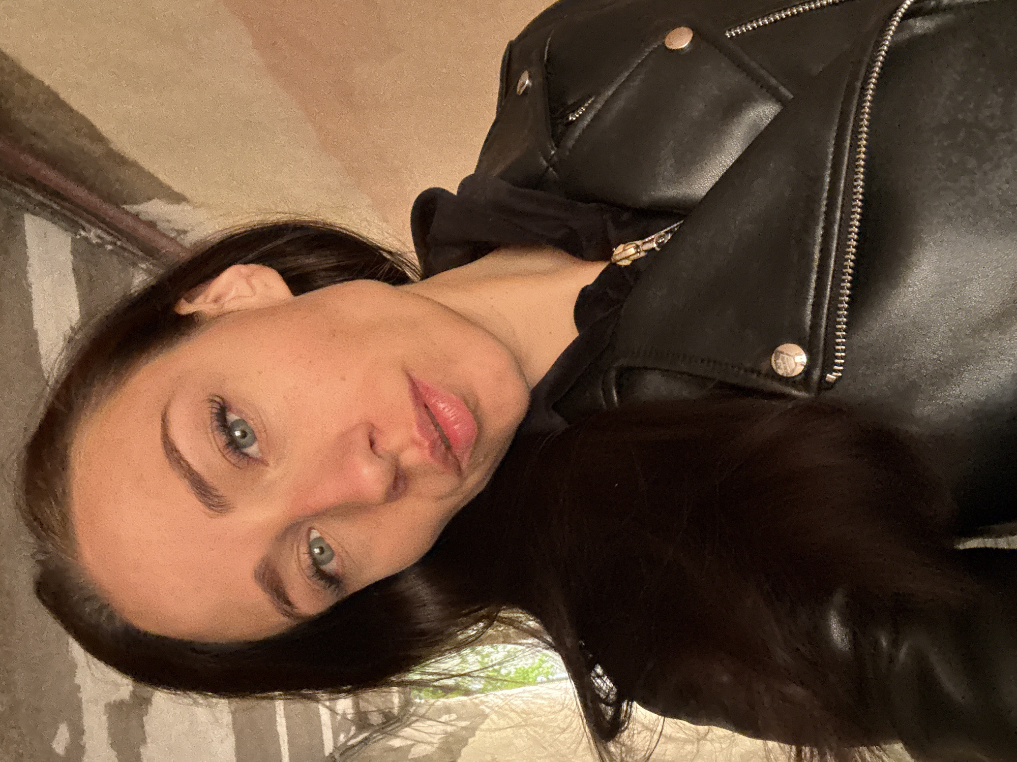A young Caucasian woman with long dark brown hair and blue eyes is taking a selfie. She is wearing a black leather jacket over a black hooded sweatshirt. The background appears to be an interior space with textured walls and some exposed piping or conduits. There is a window to the left of the frame, with greenery visible outside. The overall lighting suggests an indoor environment with some natural light coming from the window. The woman is looking directly at the camera with a neutral expression. The photo was taken in Saint Petersburg, Russia.