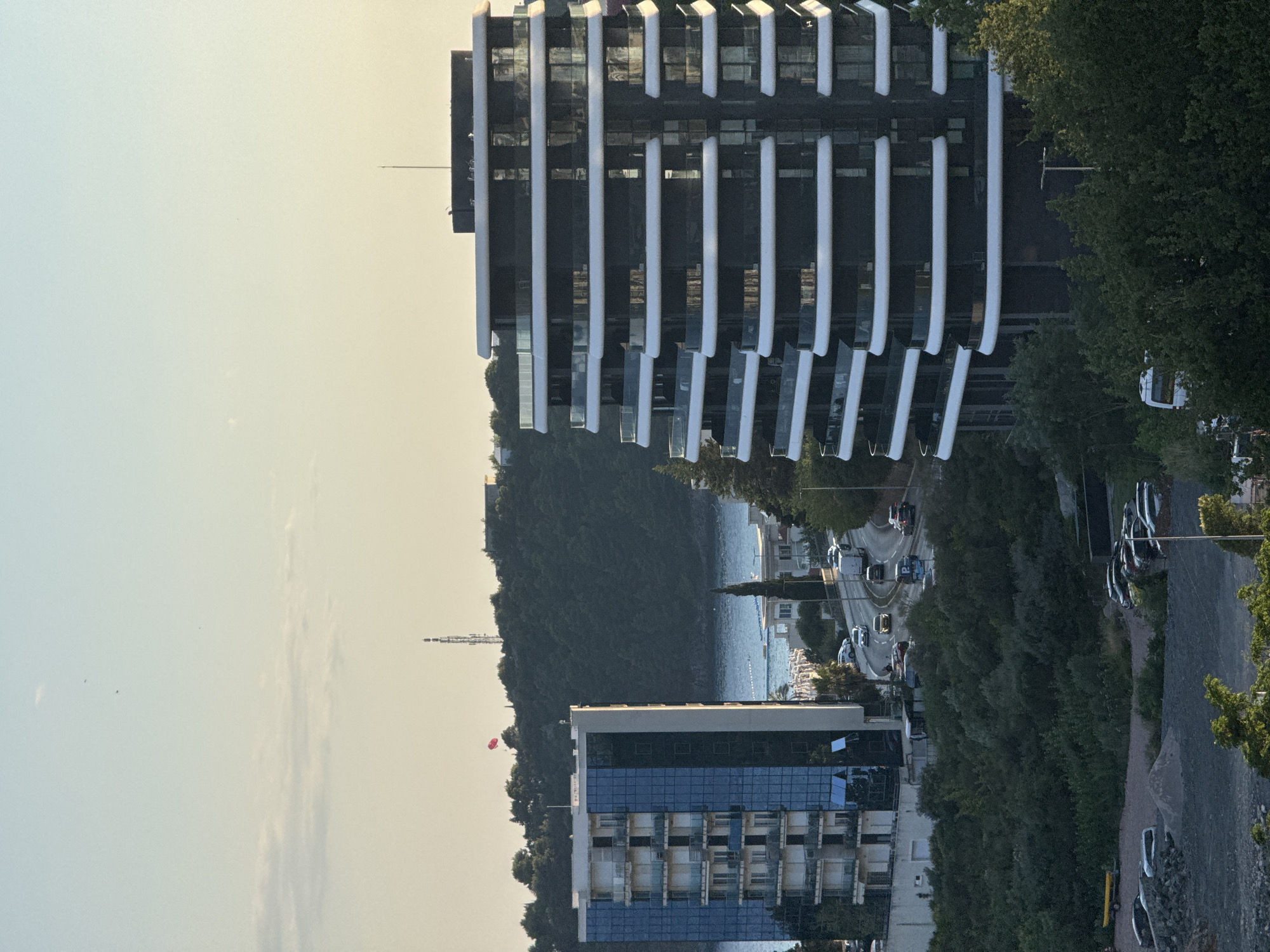 The image depicts an aerial view of Boreti, Montenegro, showcasing modern architecture against a backdrop of lush greenery and the Adriatic Sea. Dominating the foreground is a distinctive, multi-story building with a curved facade, characterized by horizontal bands of dark glass and white balconies. To its left, a rectangular building with blue glass windows stands tall, featuring a similar balcony structure.
A road curves through the scene, with several vehicles, including cars and a truck, visible in motion. The sea, a tranquil blue, is visible in the middle ground, dotted with white buoys or markers. Beyond the water, a verdant hillside rises, topped with trees and a telecommunications tower. A red parachute is visible in the sky above the trees, suggesting recreational activity.
The lower portion of the image reveals a gravel lot with several parked cars and a yellow trailer. Dense green foliage covers the landscape, softening the urban elements and integrating them into the natural environment. The sky is a pale gradient of blue and yellow, indicating either dawn or dusk.
