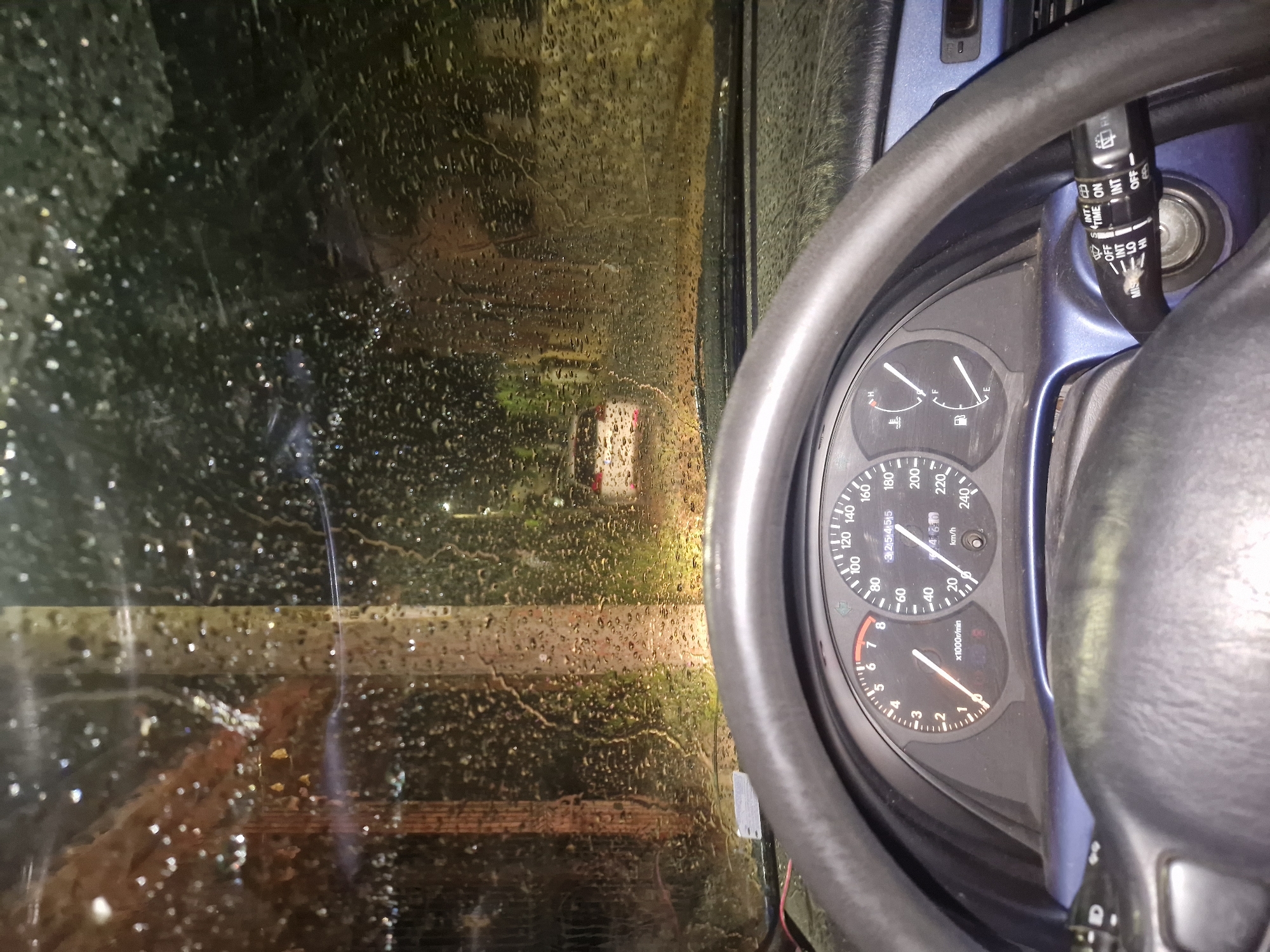 The image captures an interior view from a car, likely in Rila, Bulgaria, at night or in very low light conditions, during or immediately after rainfall. The primary subject is a car's dashboard and a rain-soaked windshield.
On the right side of the frame, the black steering wheel and a section of the dashboard are prominent. The instrument cluster features a speedometer marked up to 240 km/h and a tachometer reaching 8 x1000r/min, both indicating zero, suggesting the car is stationary or off. A digital display shows "325145.5" and "460," likely representing an odometer and trip meter respectively. Fuel and temperature gauges are also visible. To the right of the steering wheel, a wiper control stalk is seen, featuring various settings such as "INT" (intermittent), "OFF", "ON", "HI" (high speed), "LO" (low speed), "MIN", "TIME", and "MIST," along with corresponding icons. The car's interior trim around the dashboard and steering column appears blue.
The left side of the image is dominated by the wet windshield, covered with countless individual raindrops and streaks of water. Through the blurred, rain-distorted glass, a dark, indistinct exterior scene is visible. A bright, yellowish light source in the distance casts a significant glow, indicating the presence of artificial illumination like streetlights or another vehicle's headlights. Other fainter, diffused lights contribute to the dark, wet ambiance outside the vehicle. The scene conveys a sense of being sheltered inside the car while observing a rainy environment.