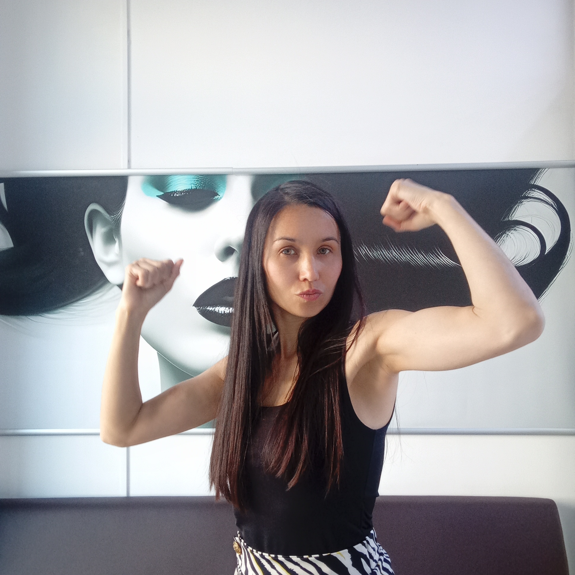 A young woman with long, dark hair is posing indoors, flexing her biceps. She is wearing a black tank top and a black and white patterned skirt. Behind her is a large, modern artwork featuring a stylized face with bold makeup, set against a white wall. The woman's arms are raised, with her fists clenched, displaying her biceps. The photo is taken from a medium distance, showing her from the waist up. The setting appears to be a contemporary interior, possibly a waiting area or office space. There is no information about the image being taken in Izhevsk, Russia.