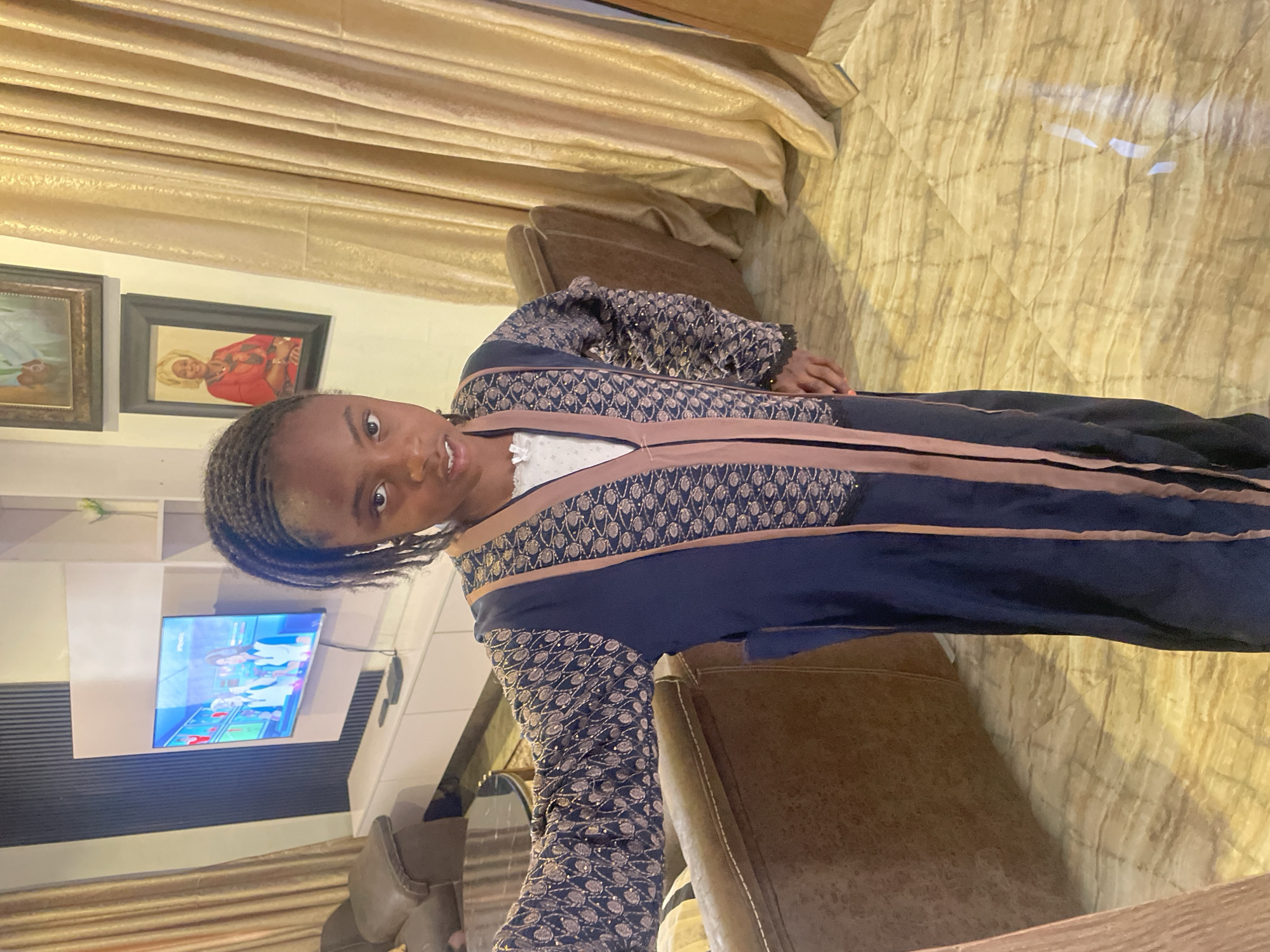 A young girl is indoors. She is wearing a long, ornate, navy blue dress with gold trim. The room has cream-colored curtains and a beige tiled floor. There are two framed portraits on the wall and a TV with several people on it. The girl is seated on a brown chair. The location is Jos, Nigeria.